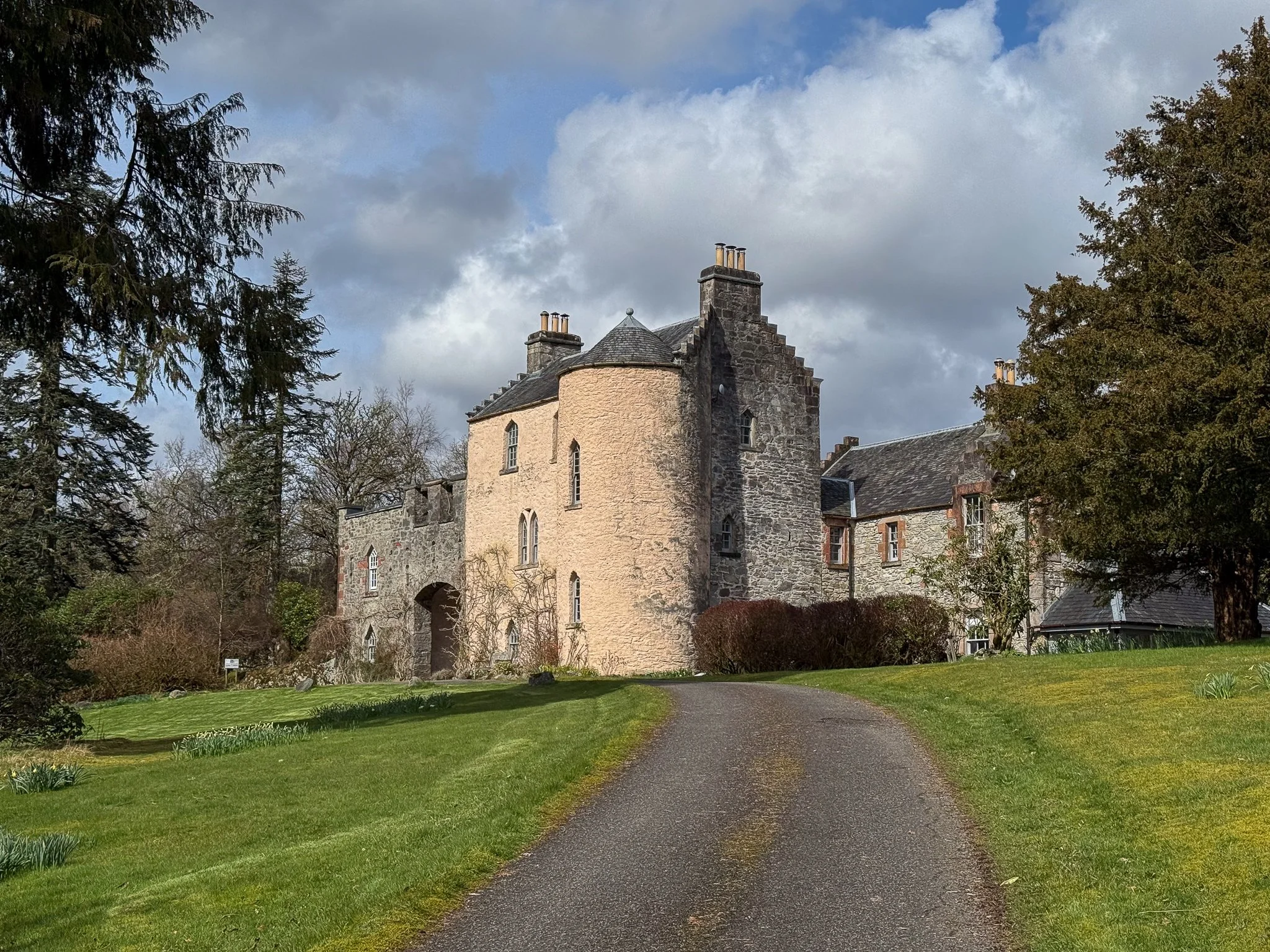 A historic stone castle with a curved tower and multiple chimneys, surrounded by a well-maintained lawn and trees, under a partly cloudy sky.