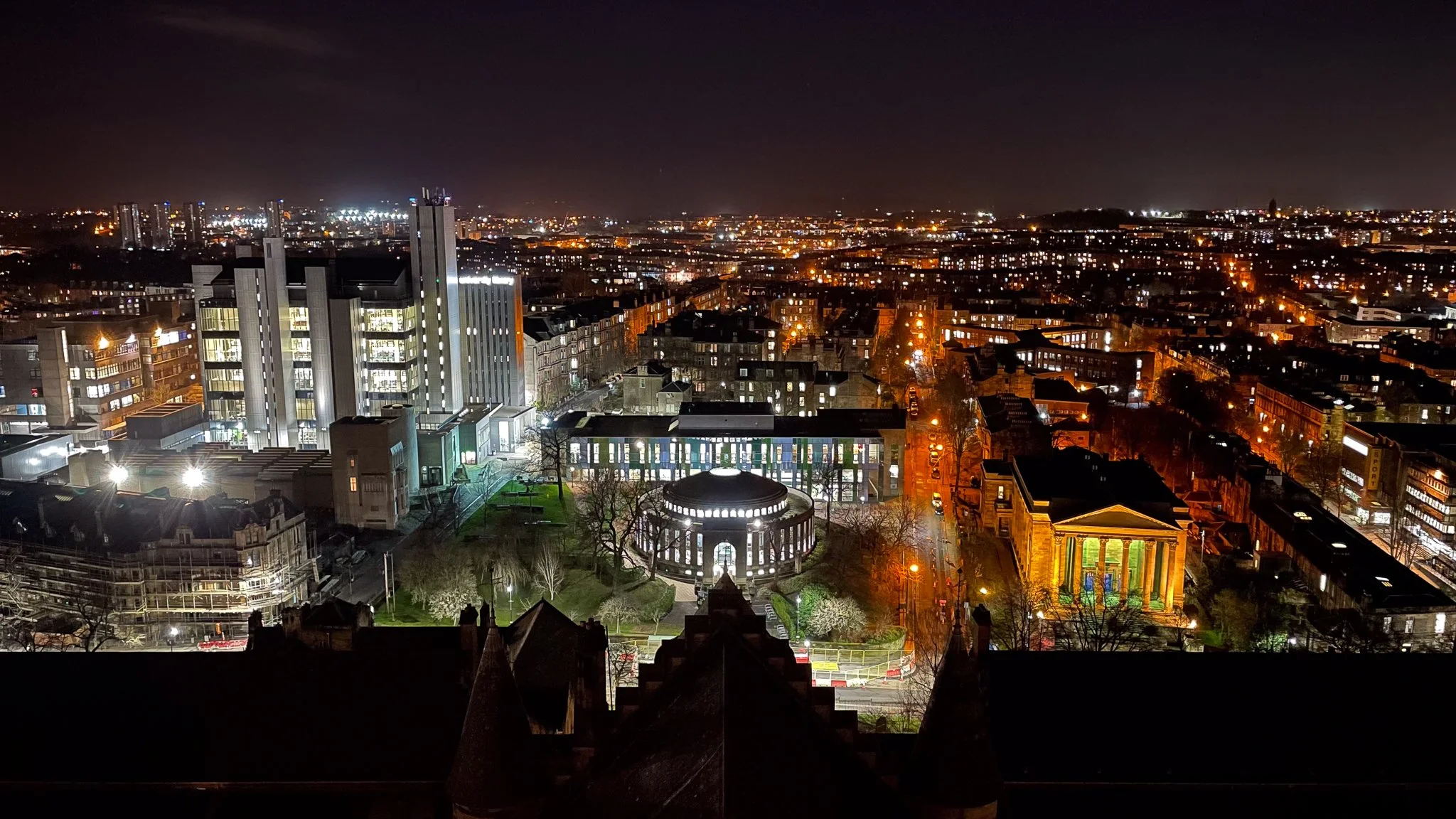 Nighttime panoramic city view with illuminated buildings, streets, and landmarks, including a historic round building and a neoclassical temple-style structure.