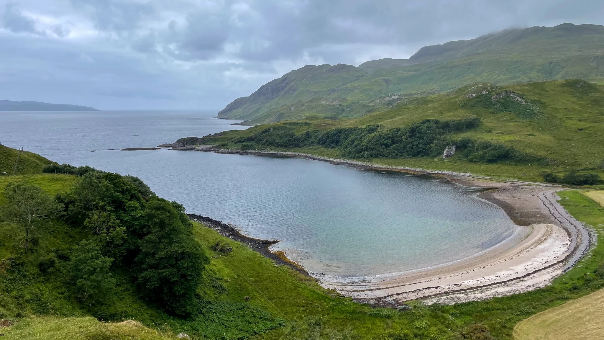 A scenic view of a bay with a sandy beach, green hills, and mountains in the background under cloudy skies.