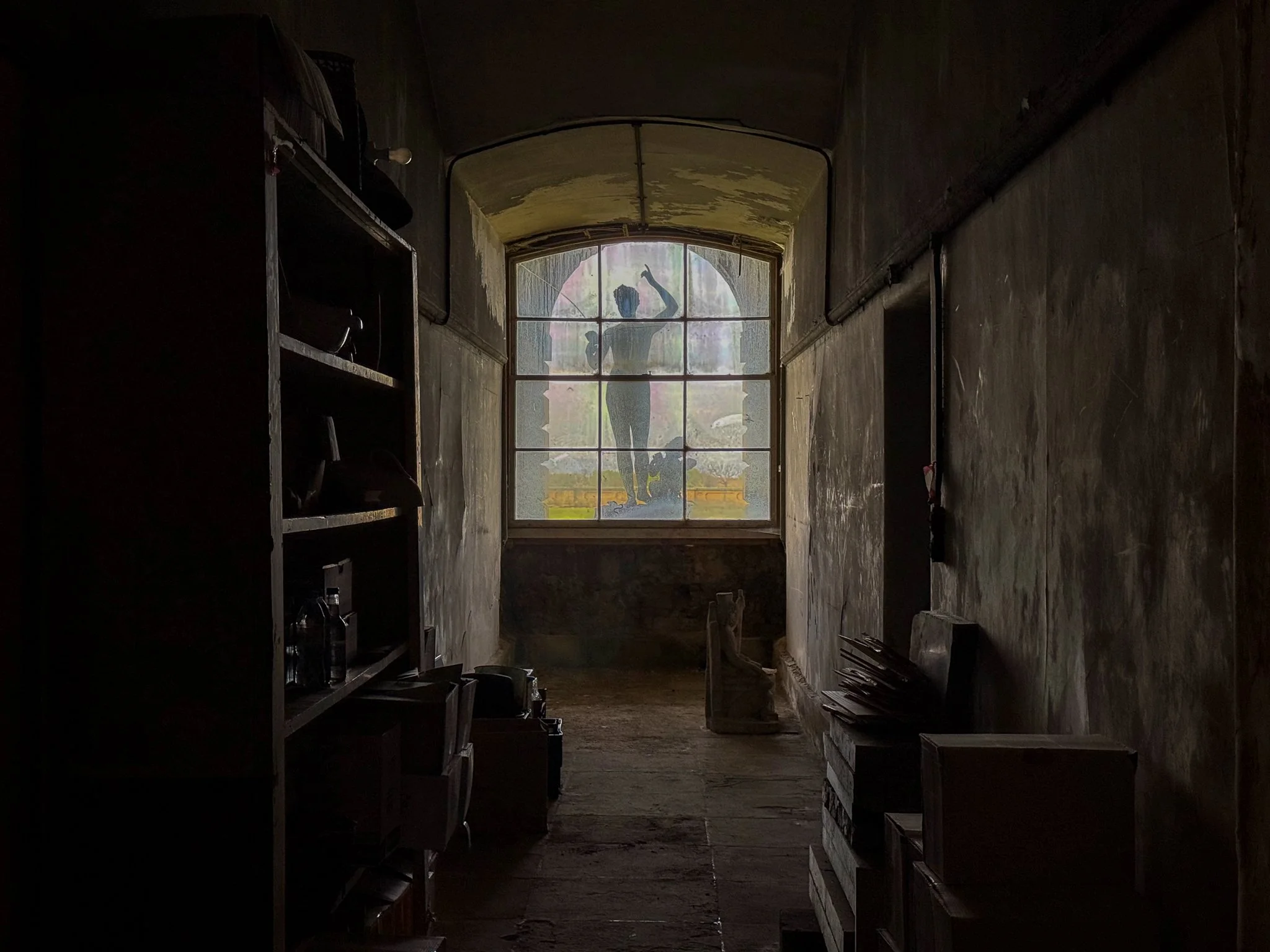 Dark and abandoned hallway with a large window at the end, featuring a silhouette of a woman painting on the glass, with some shelves and furniture along the sides.