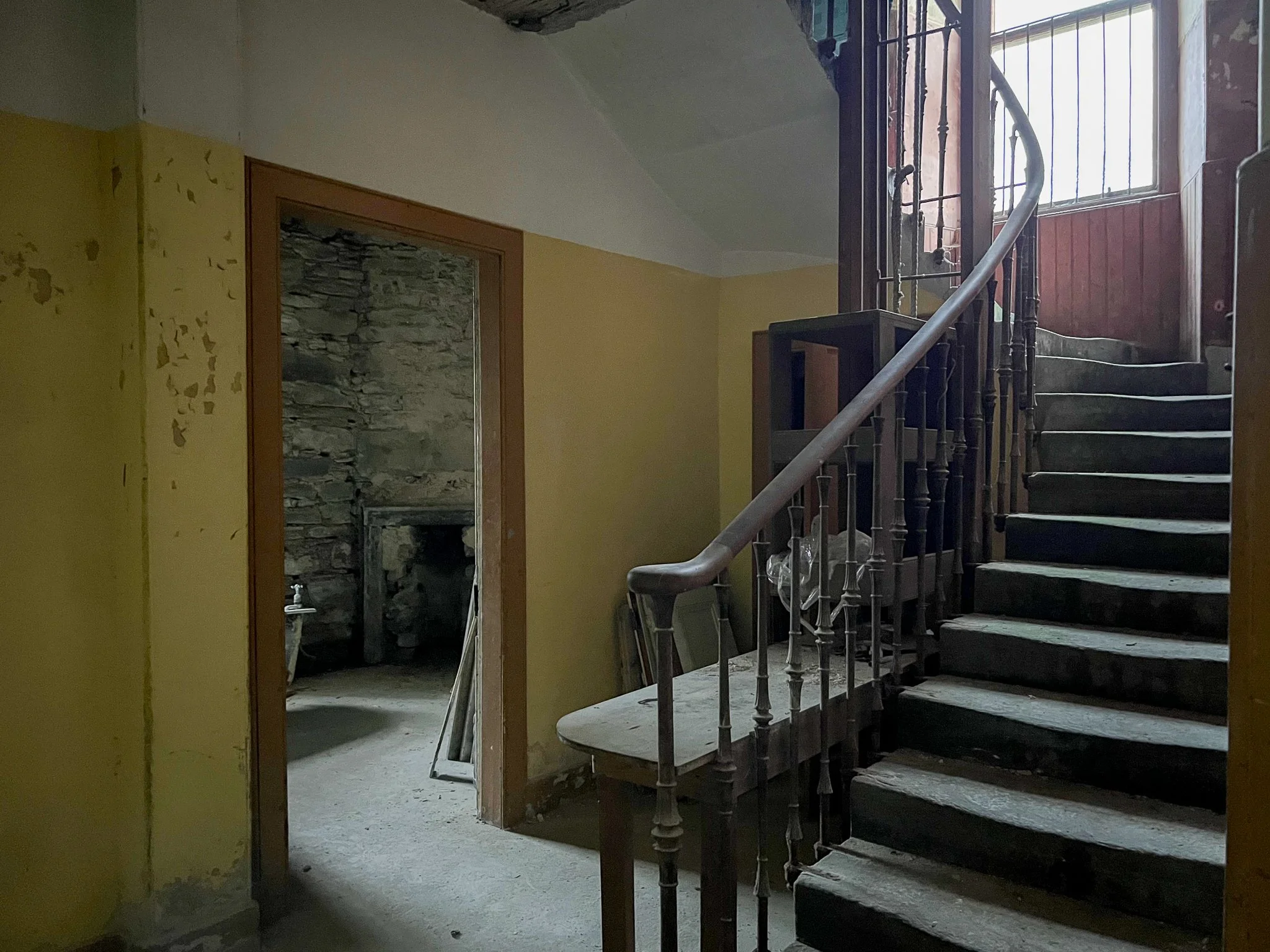 A dingy, abandoned interior stairway with a wooden handrail and access to a small, dark room on the left. The staircase has worn, old steps and is illuminated by natural light from a window at the top.
