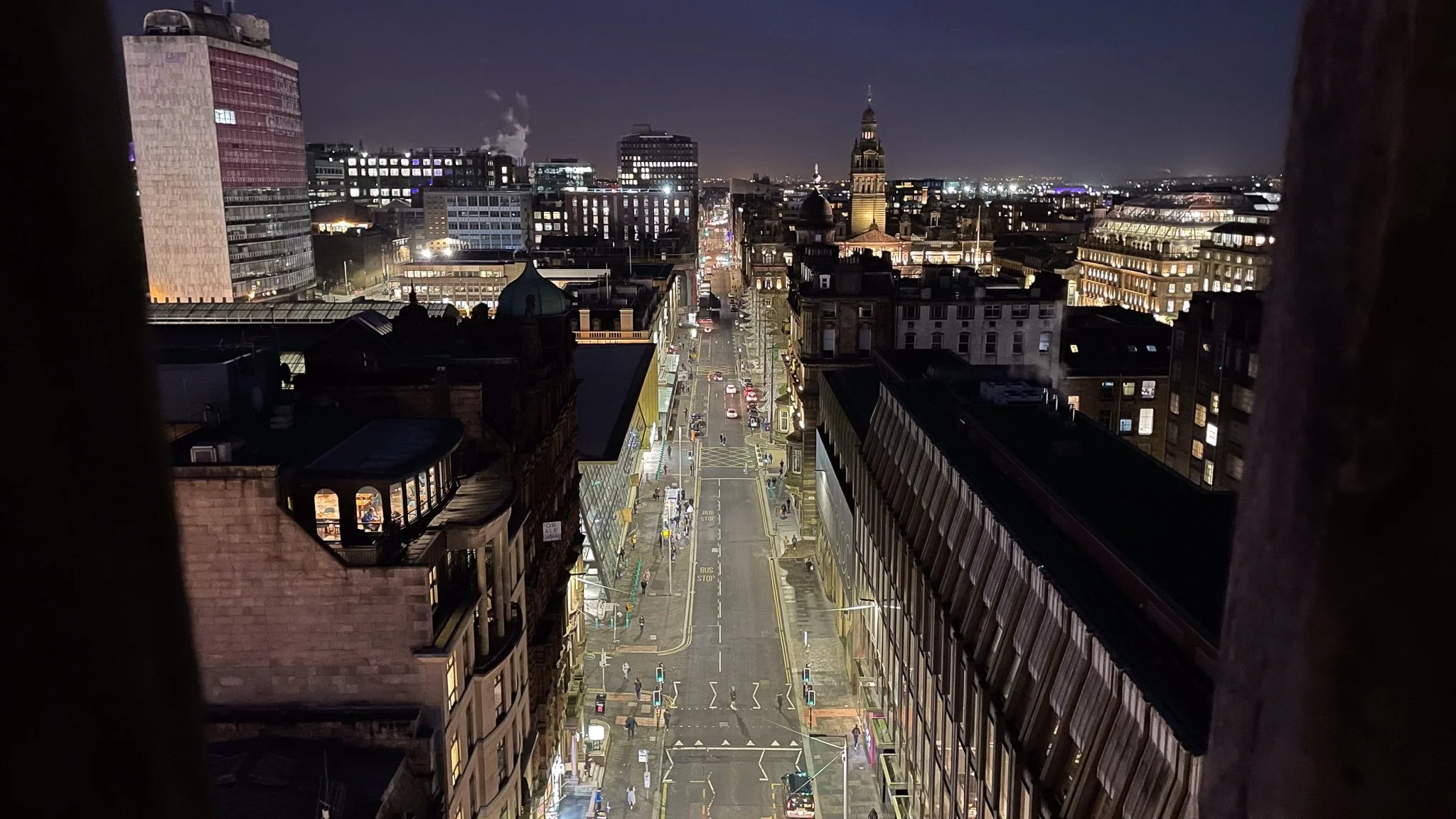 Nighttime city street view with tall buildings, illuminated sidewalks, and a distant clock tower in the background.