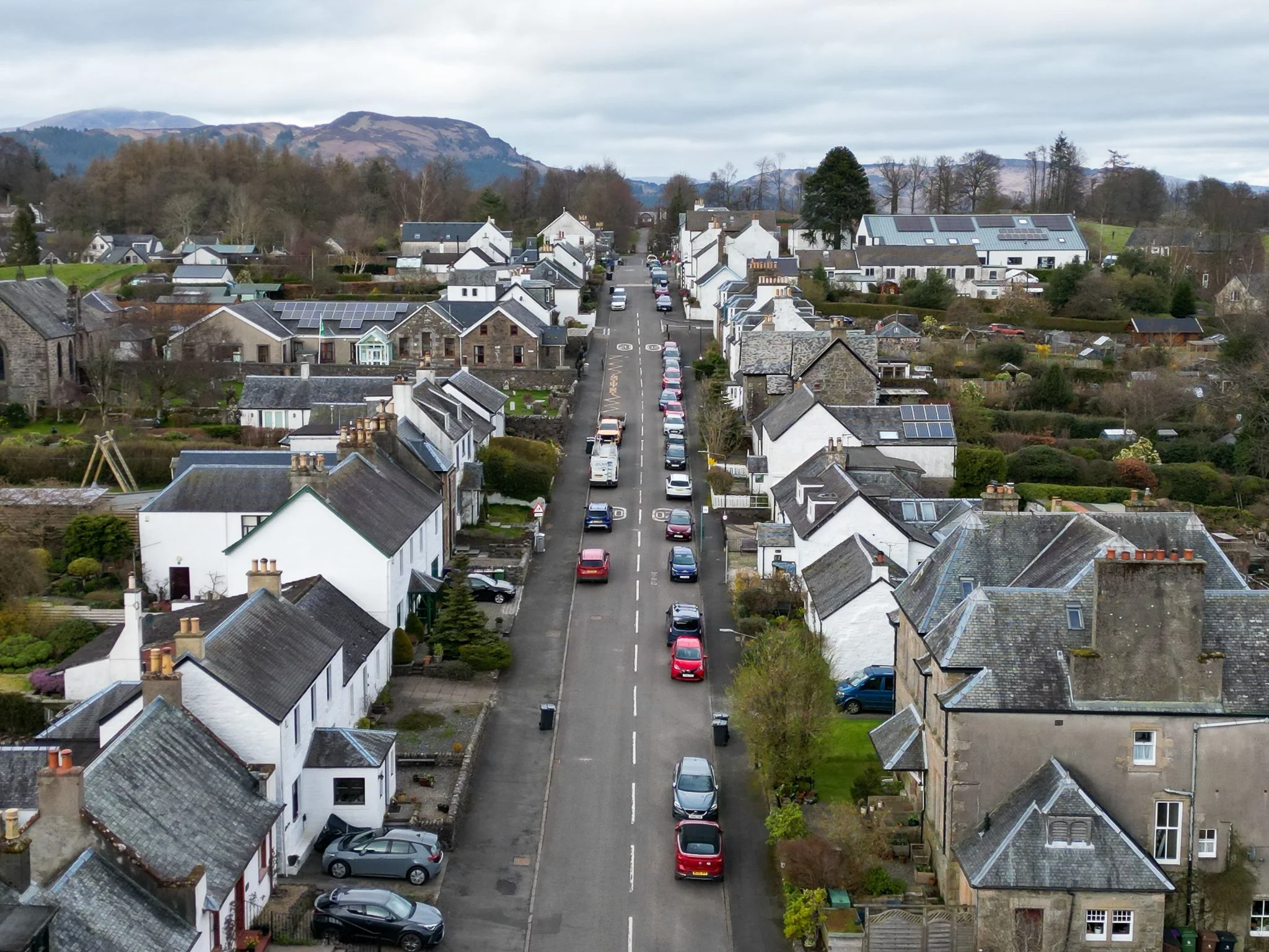 A small town street with parked cars on both sides, surrounded by houses with slate roofs, some with solar panels, and rolling hills in the background.