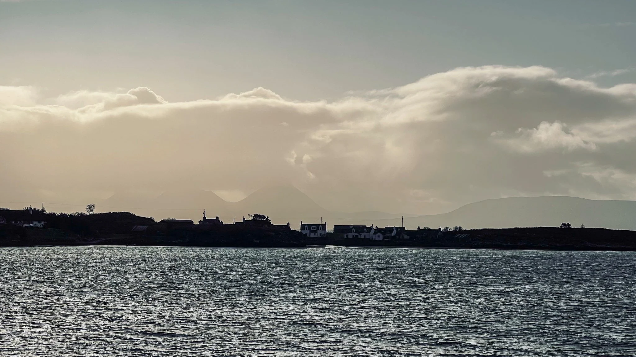 A body of water in the foreground with a shoreline featuring houses and trees, and hills in the background under a partly cloudy sky.