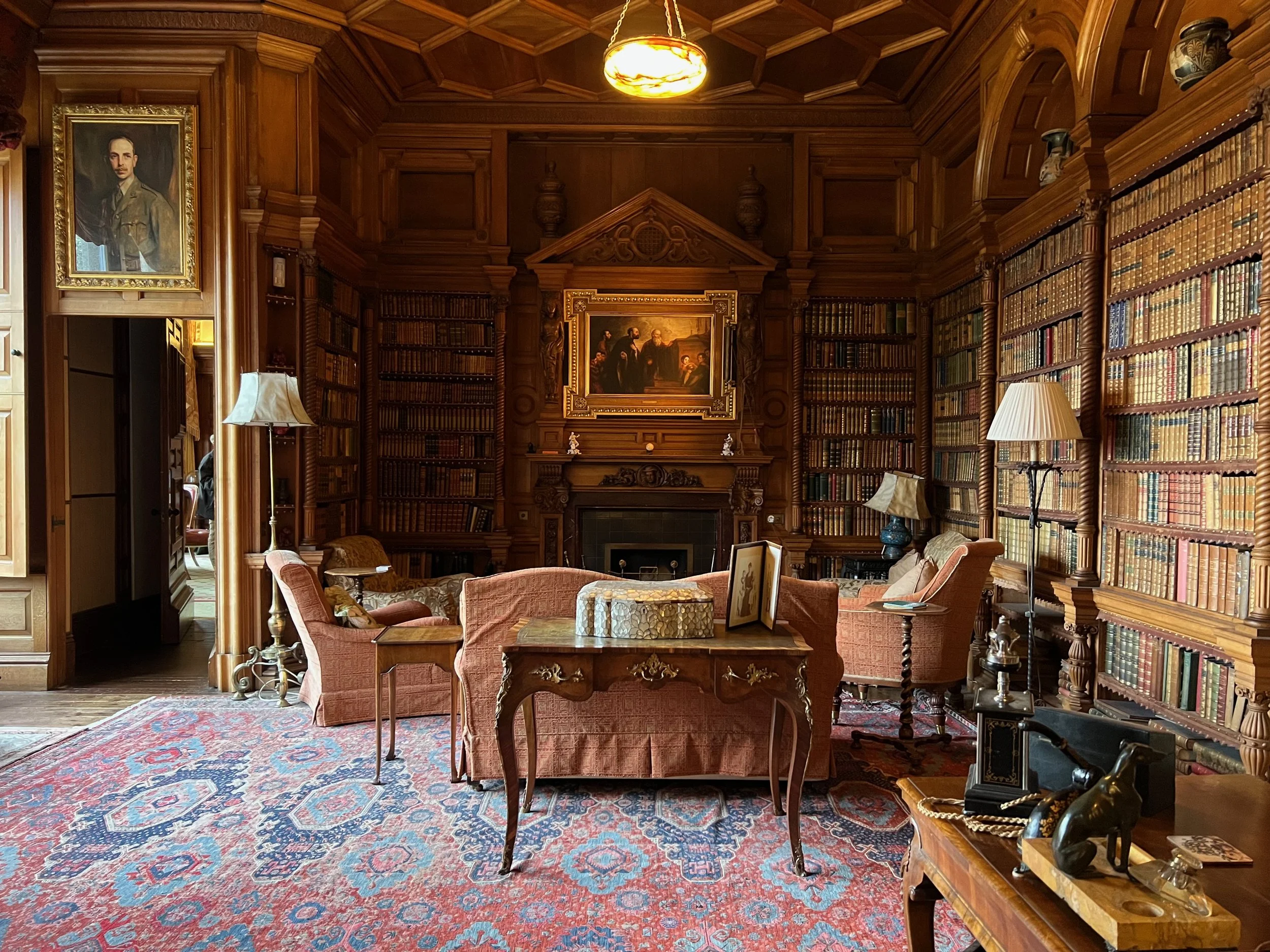 A classic, elegant library room with wood-paneled walls and floor-to-ceiling bookshelves filled with leather-bound books. There are upholstered armchairs, a patterned area rug, lamps, a fireplace, and a large framed painting above the mantle. A portr