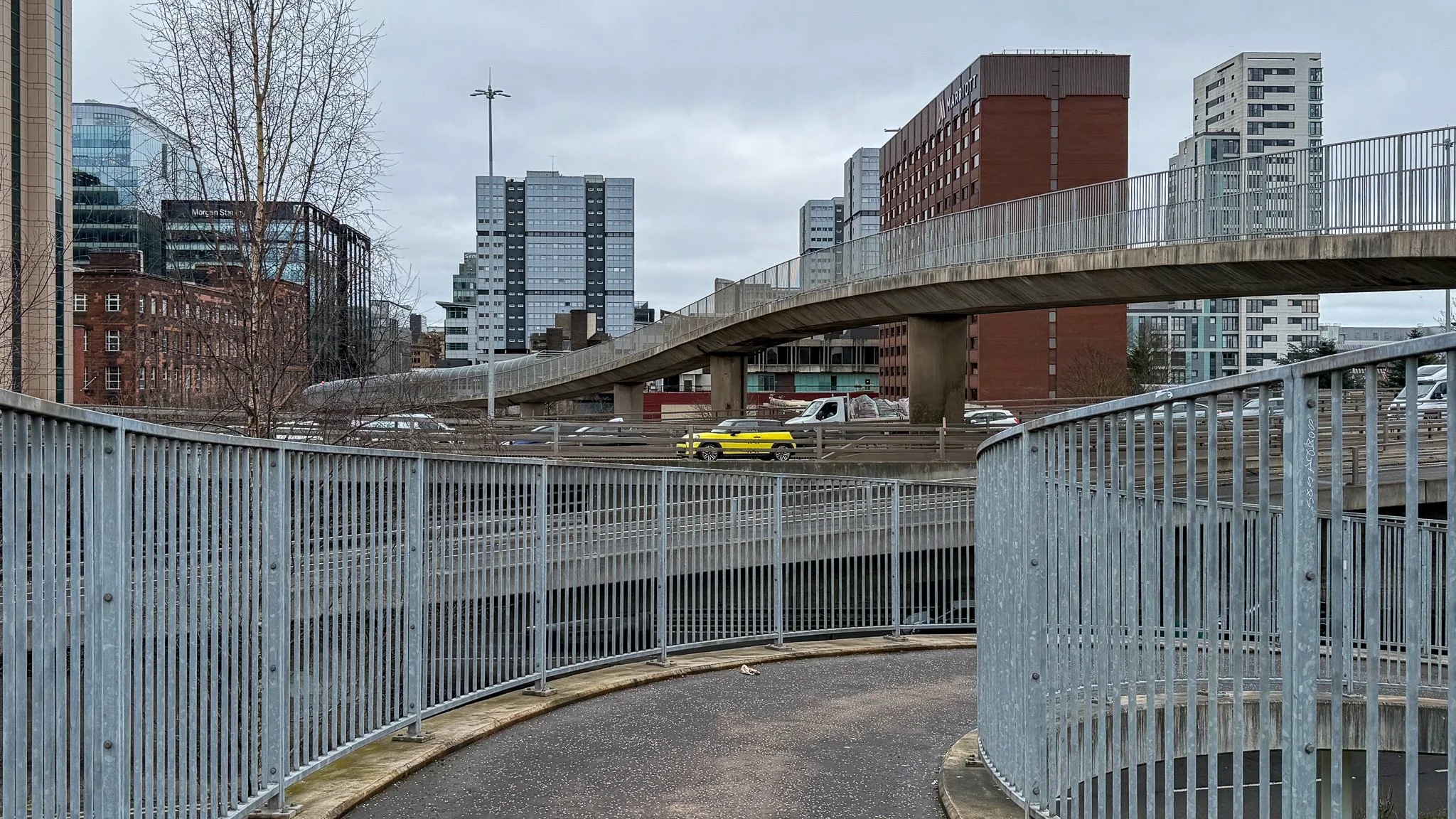 Urban scene with a curved metal pedestrian walkway and fencing, multiple high-rise buildings, parked cars, and cloudy sky.