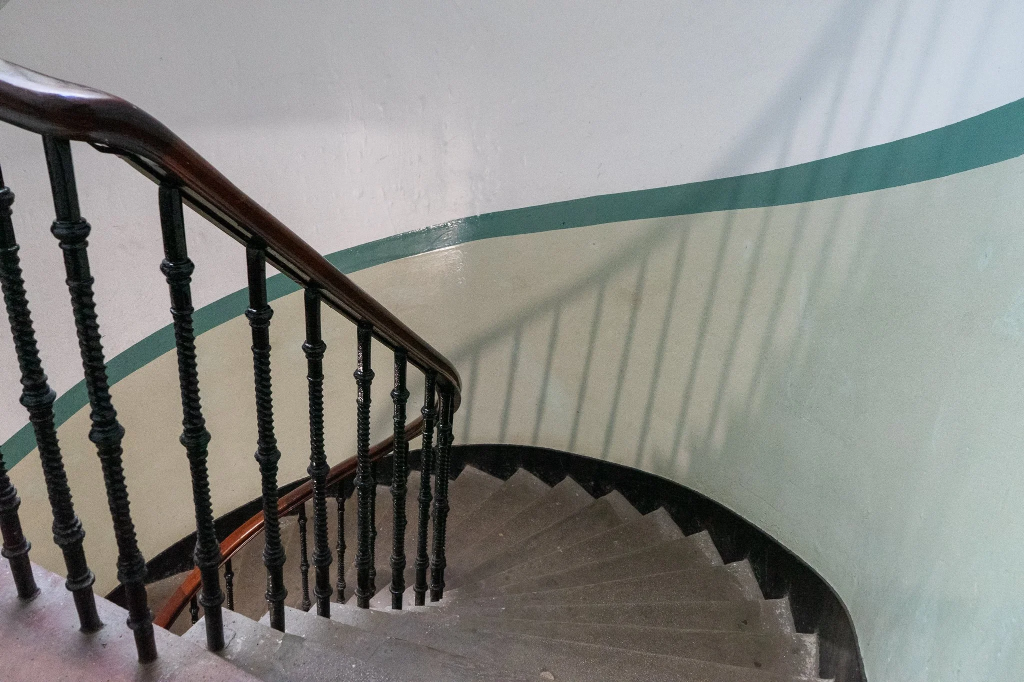 A spiral staircase with black metal balusters and a wooden handrail, casting shadows on a curved light green wall with a blue border.