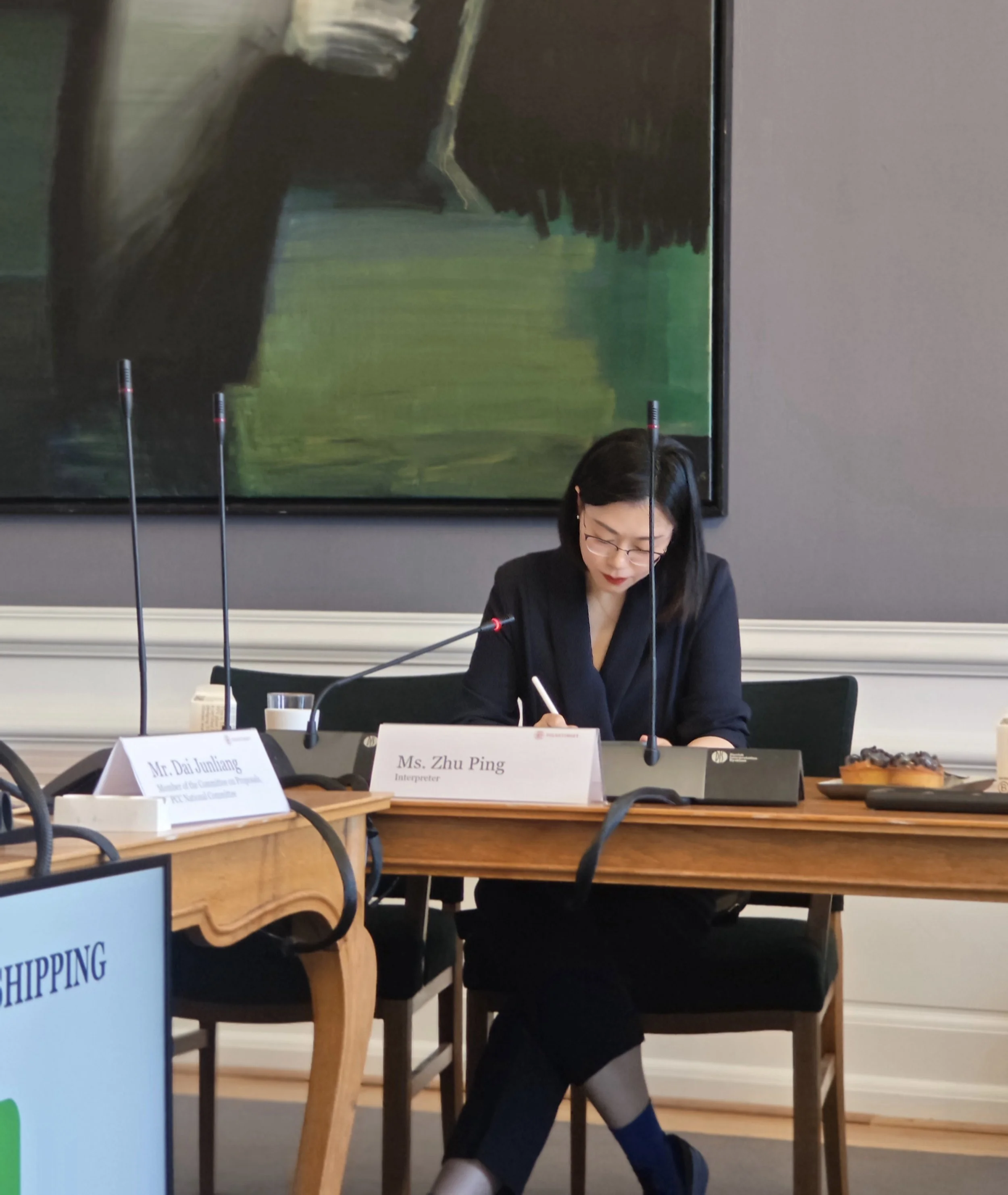 Frau sitzt am Tisch, liest Notizen, mit einem Wasser, vor zwei Mikrofonen, im Hintergrund ein großes abstraktes Gemälde an der Wand.