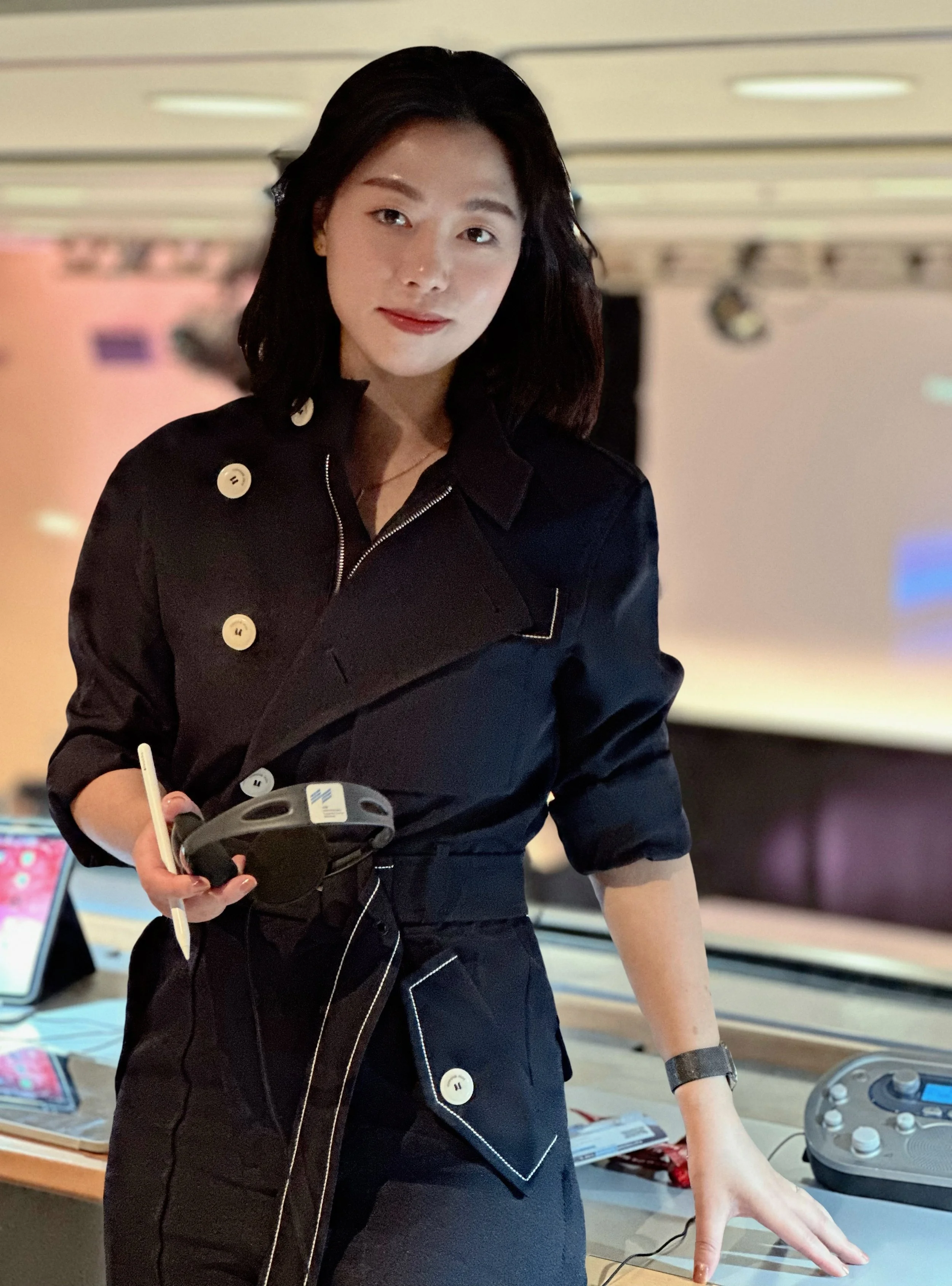 Eine junge Frau mit schwarzen Haaren trägt einen schwarzen Mantel und hält einen Druckstift in der Hand. Sie steht an einem Tisch mit elektronischem Equipment in einem Raum mit hellem, mehrstufigem Beleuchtungssystem.