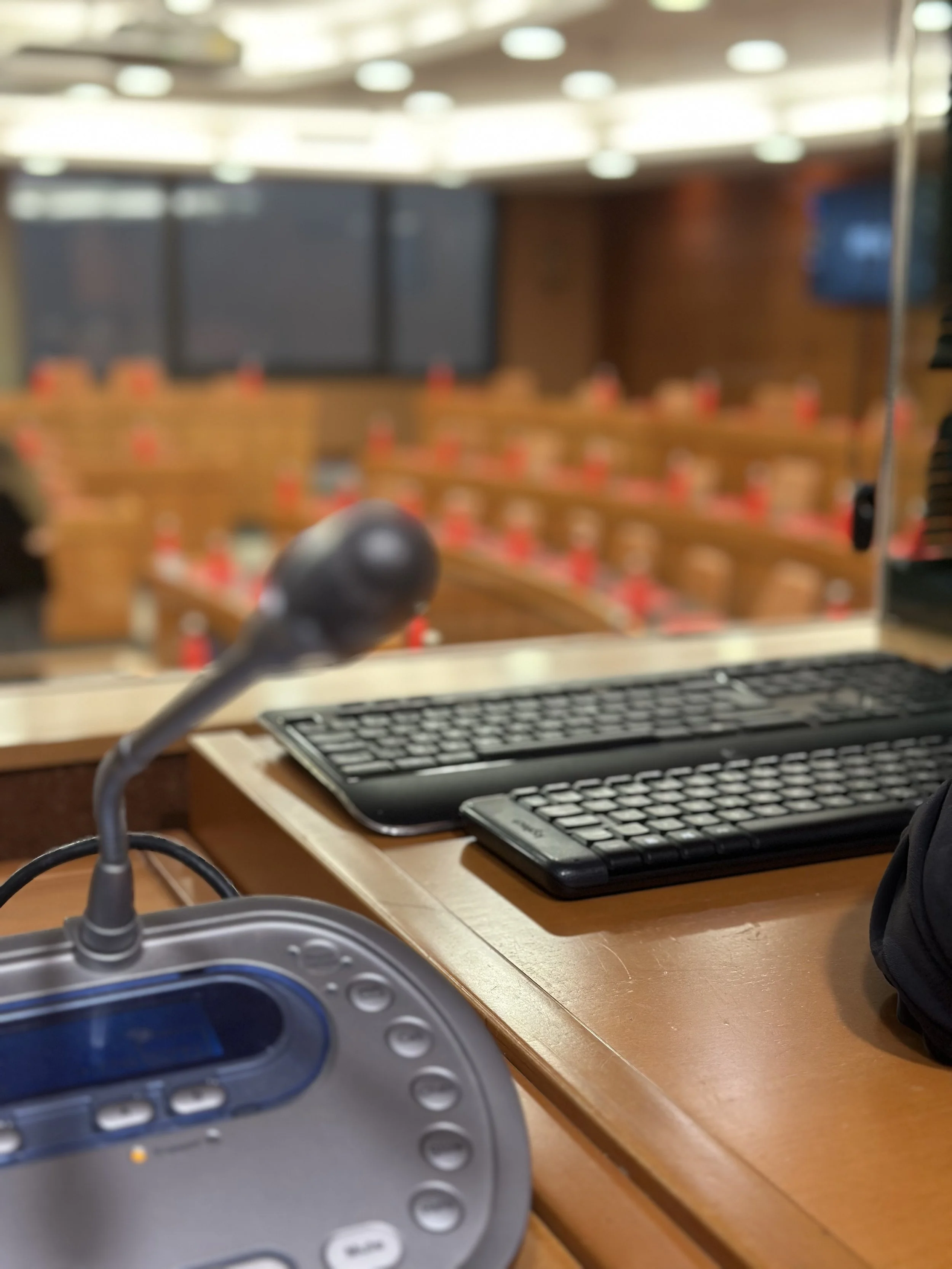 Leerer Klassenraum mit Stühlen und Tischen, die mit roten Wasserflaschen versehen sind, und einem großen Bildschirm an der Wand. Im Vordergrund befindet sich ein Rednerpult mit einem Mikrofon und zwei Tastaturen.