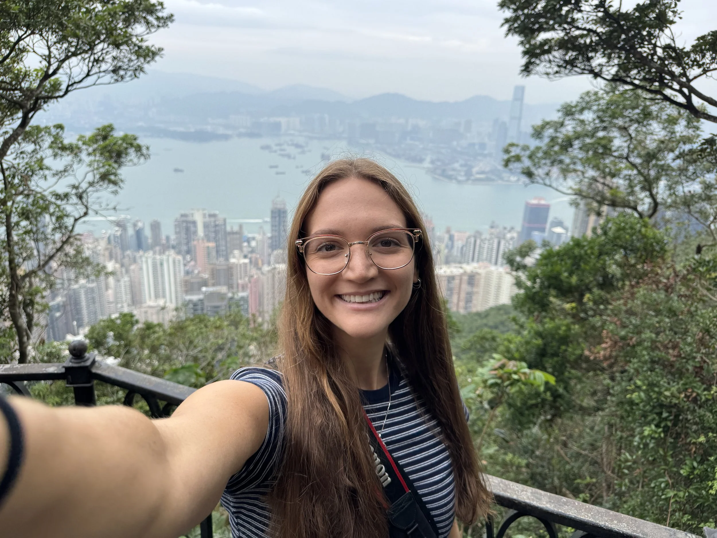 A young woman with long brown hair, glasses, and a striped shirt is smiling for a selfie at a scenic overlook with a cityscape, water, and mountains in the background. She is surrounded by green trees.