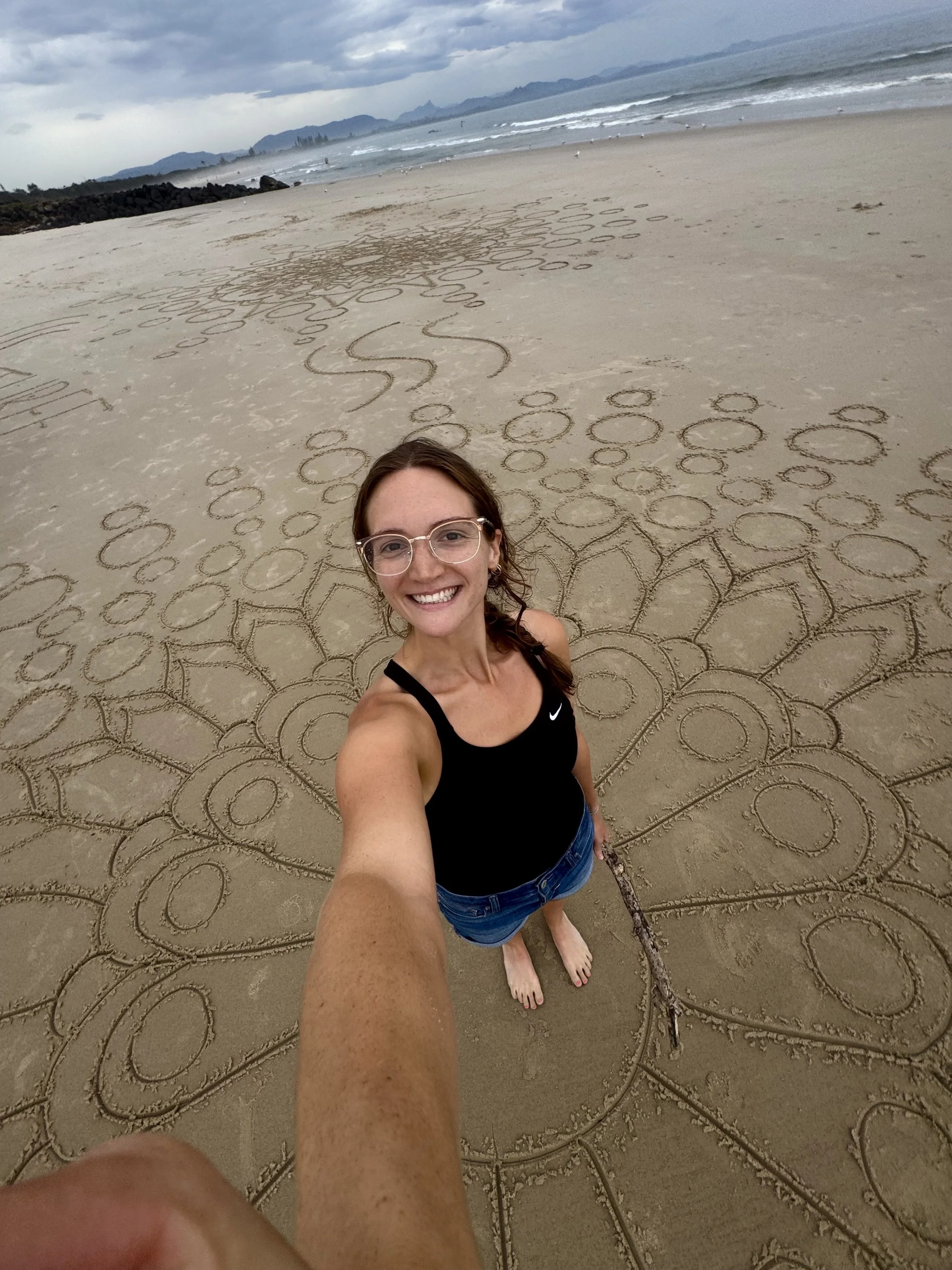 Young woman taking a selfie on a sandy beach with intricate sand art circles and patterns, ocean waves in the background under cloudy sky.
