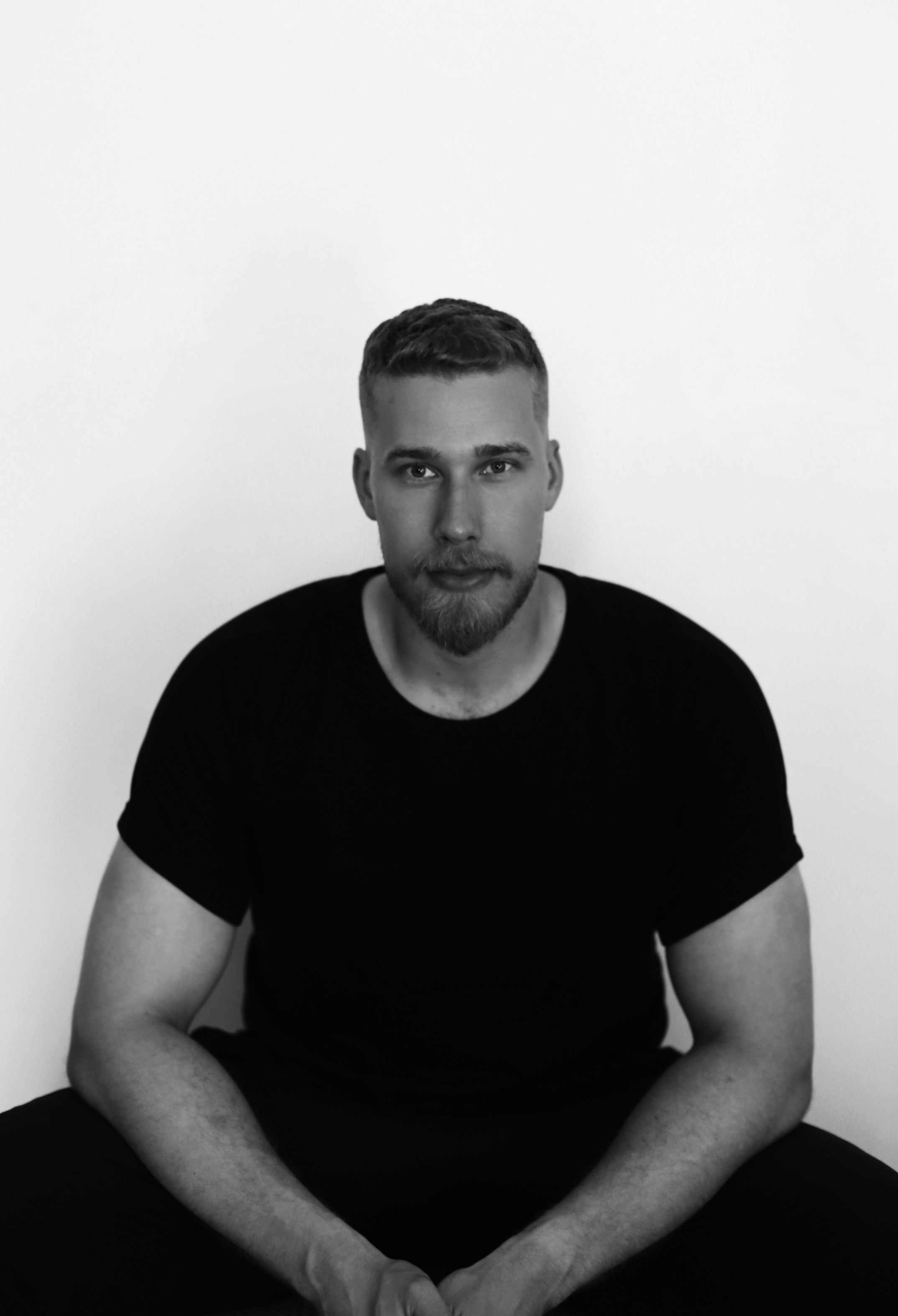 Black and white photo of a man with short hair, a beard, and wearing a black t-shirt, sitting against a plain light background.