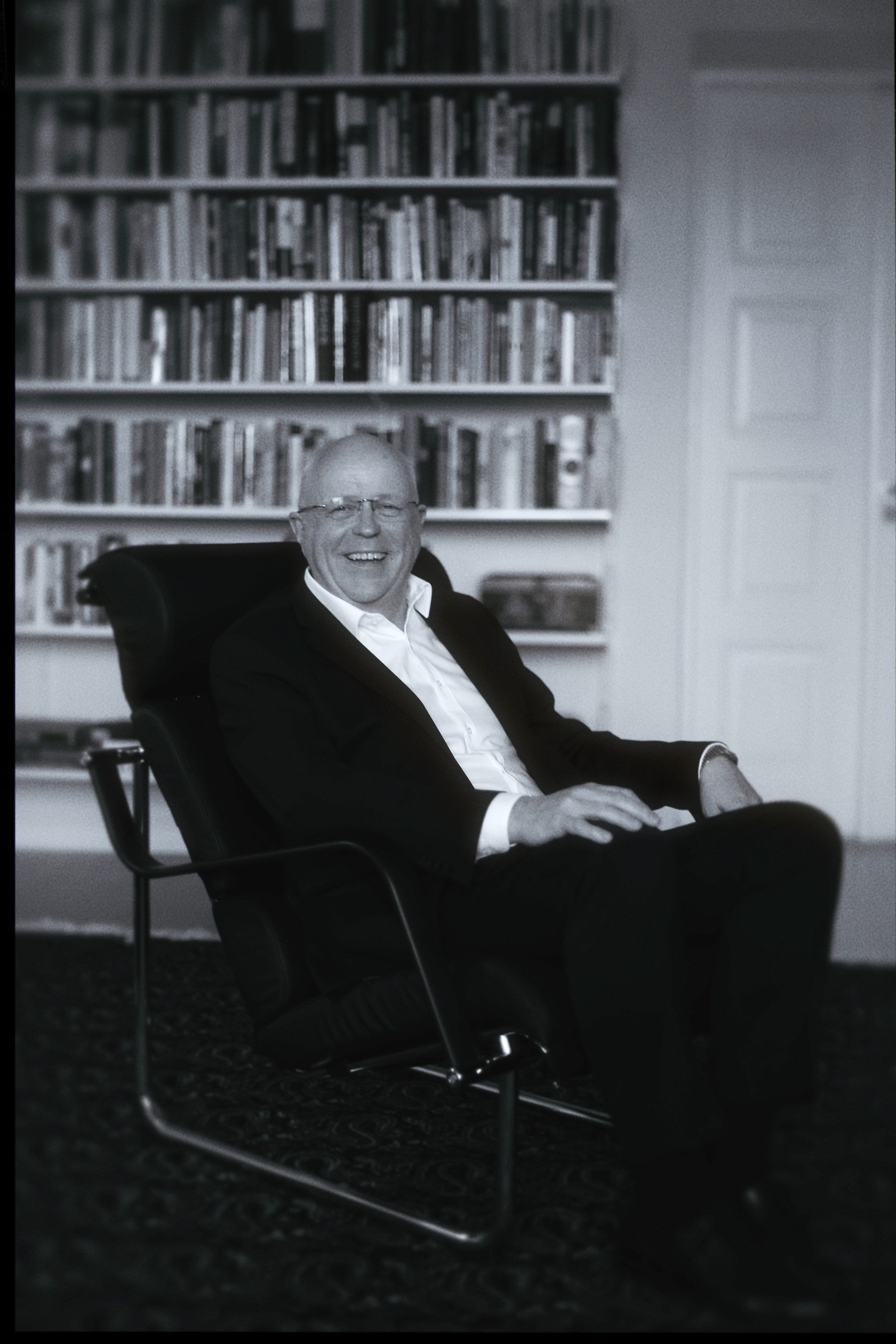 A smiling man in a suit and glasses sitting in a black chair in a room with a large bookshelf filled with books behind him.