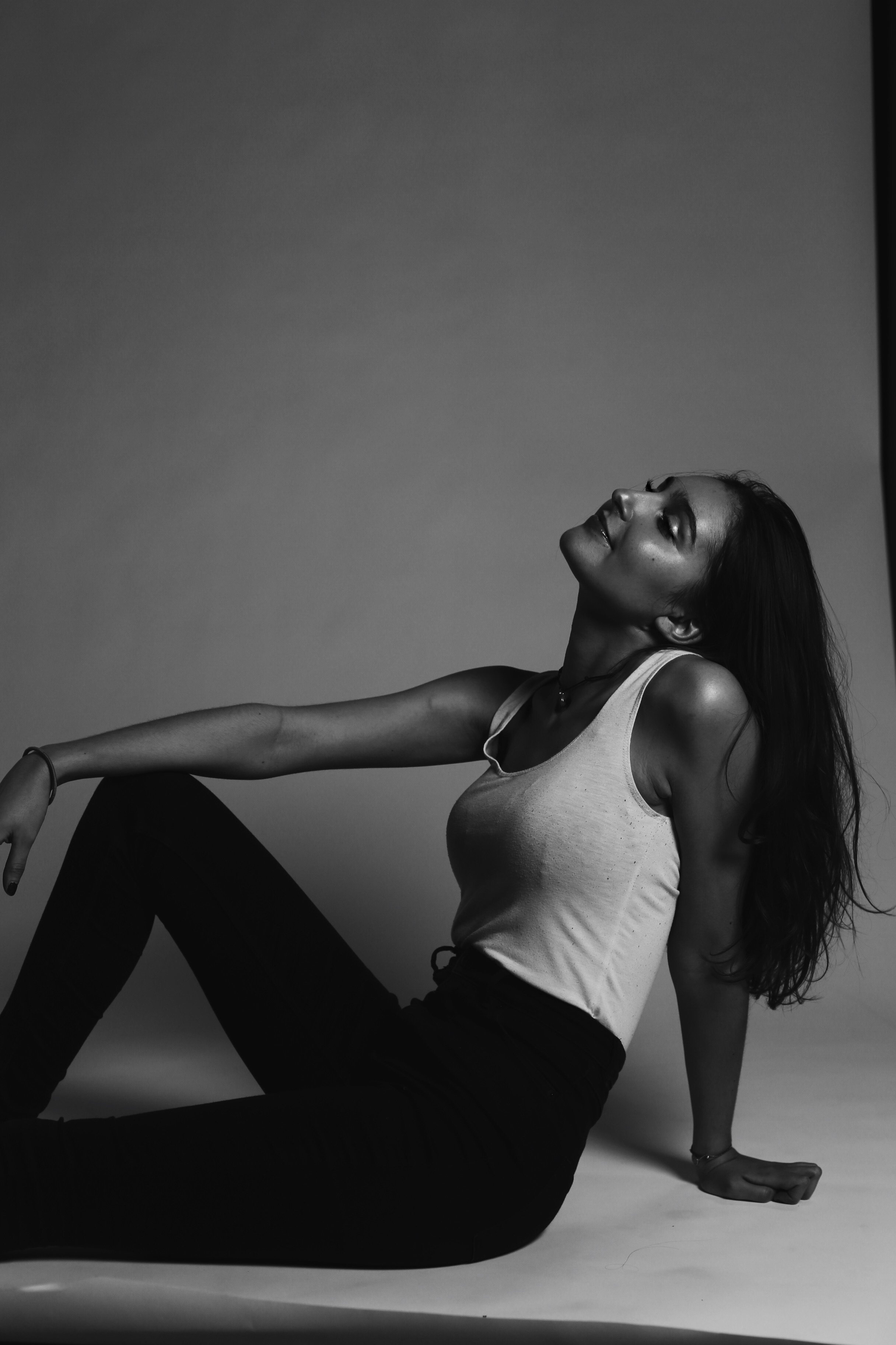 Black and white photo of a woman in a tank top and black pants sitting on the floor, leaning back on one hand with her eyes closed and a relaxed expression.