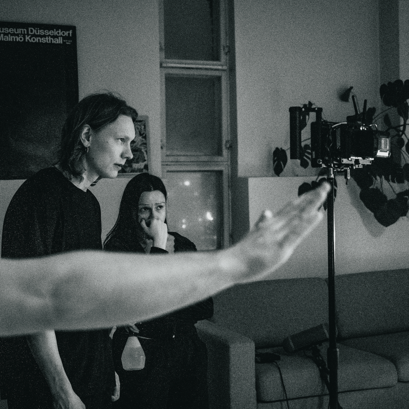 Black and white photo of two women sitting at a table during a recording or interview, with a camera on a tripod and a microphone visible. One woman has blonde hair and is looking to the left, the other woman has dark hair and is resting her face on her hand.
