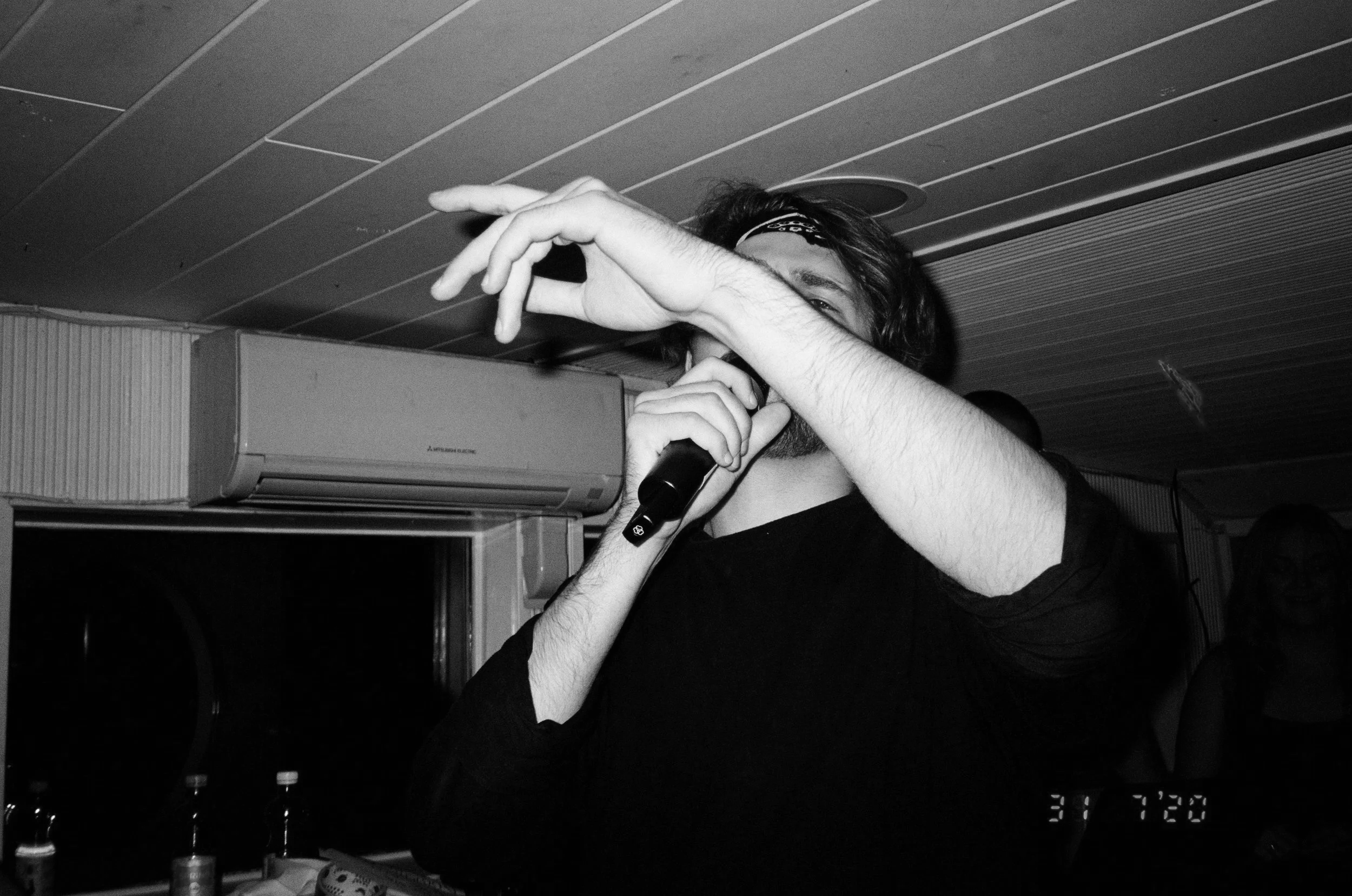 A person with long hair and a headband is holding a microphone and performing indoors, with an air conditioning unit and bottles in the background.