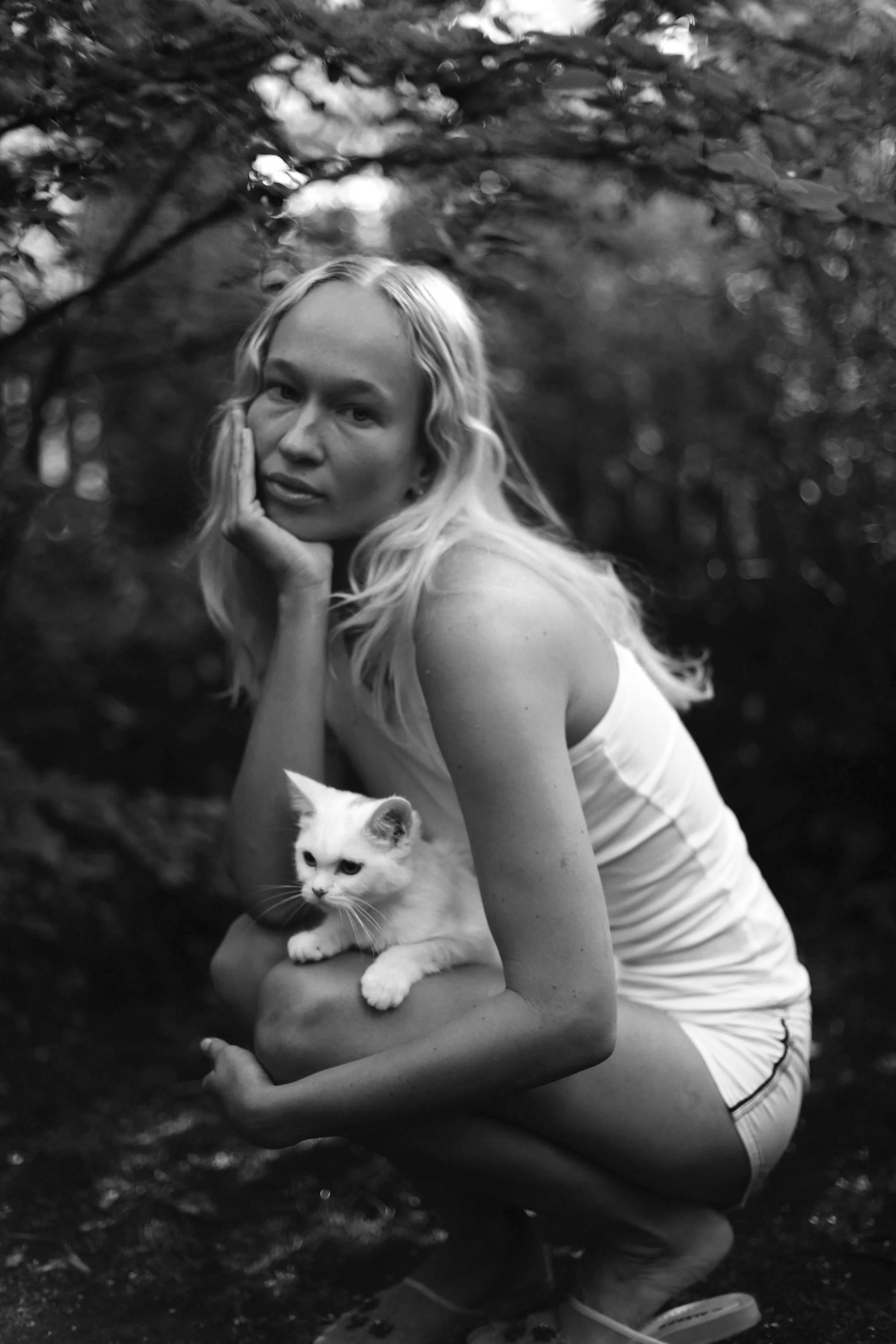 A black and white photo of a young woman with long blonde hair, wearing a white tank top and shorts, sitting outdoors with her hand resting on her cheek. She is holding a white and gray kitten in her arm, with a background of blurred trees and foliage.