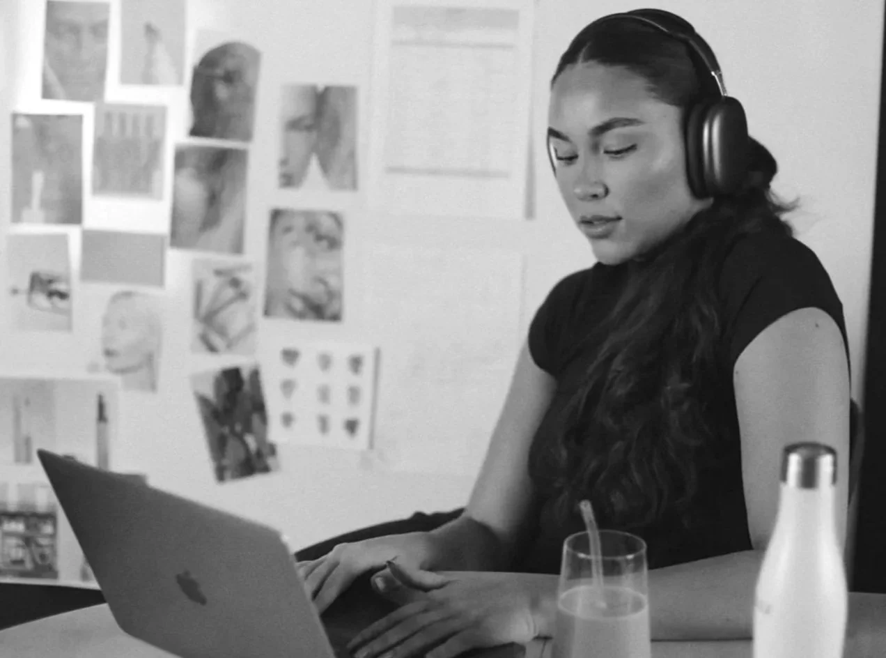 A woman with headphones working on a laptop in a room decorated with photos and artwork on the wall.