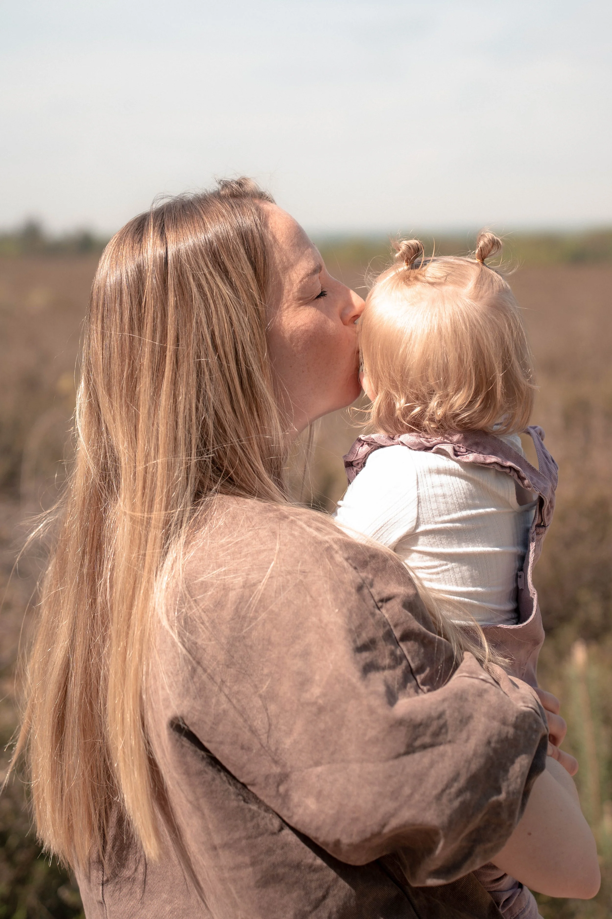 Vrouw die haar jonge dochter kust in een veld met een heldere, lichte hemel op de achtergrond.