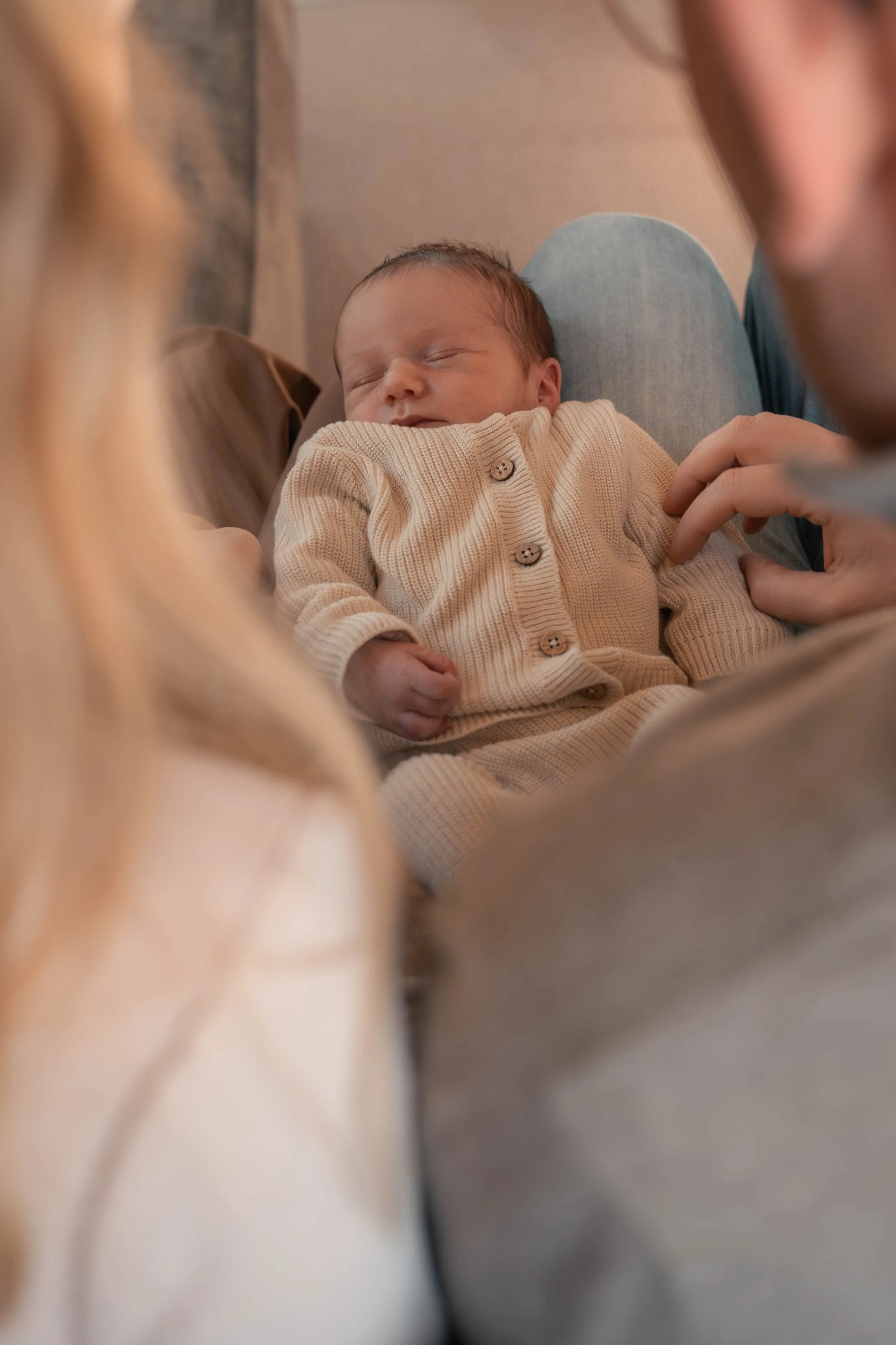 Een pasgeboren baby ligt op iemands schoot, gekleed in een beige gebreide trui met knoopjes, terwijl een volwassene naast de baby aanwezig is.