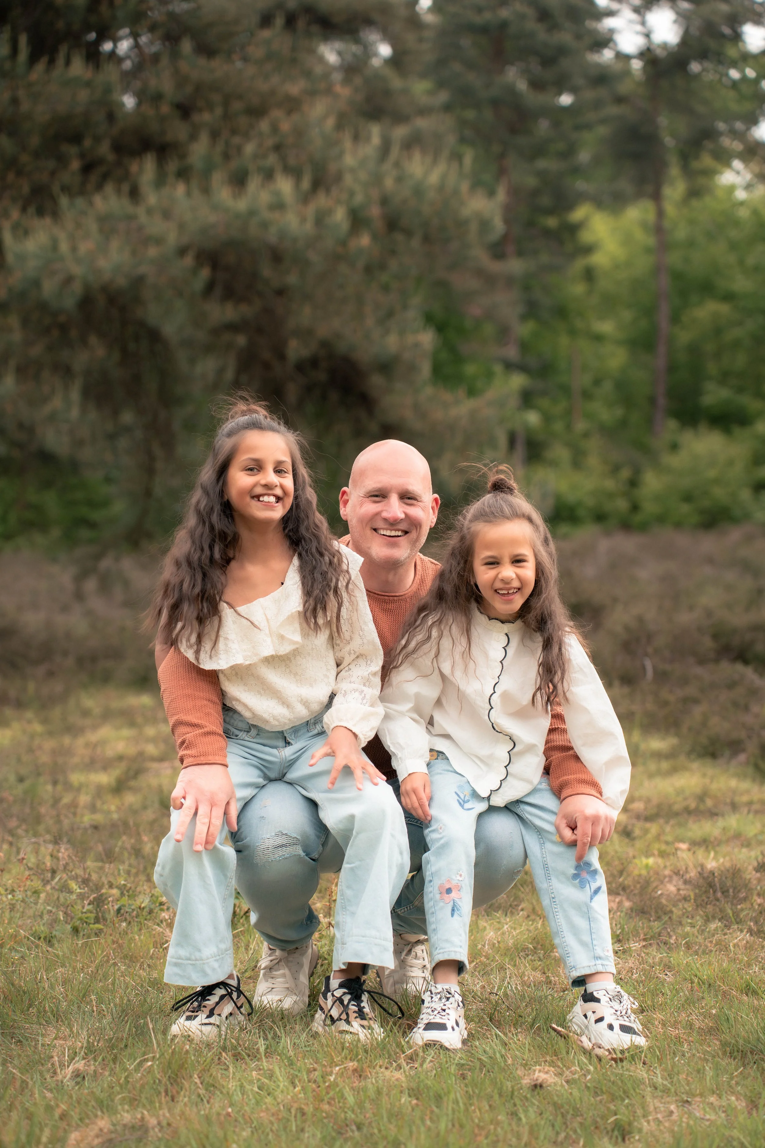 Een man en twee jonge meisjes zitten op de grond in een bosrijke omgeving met groene bomen op de achtergrond, lachend en plezier makend voor de camera.