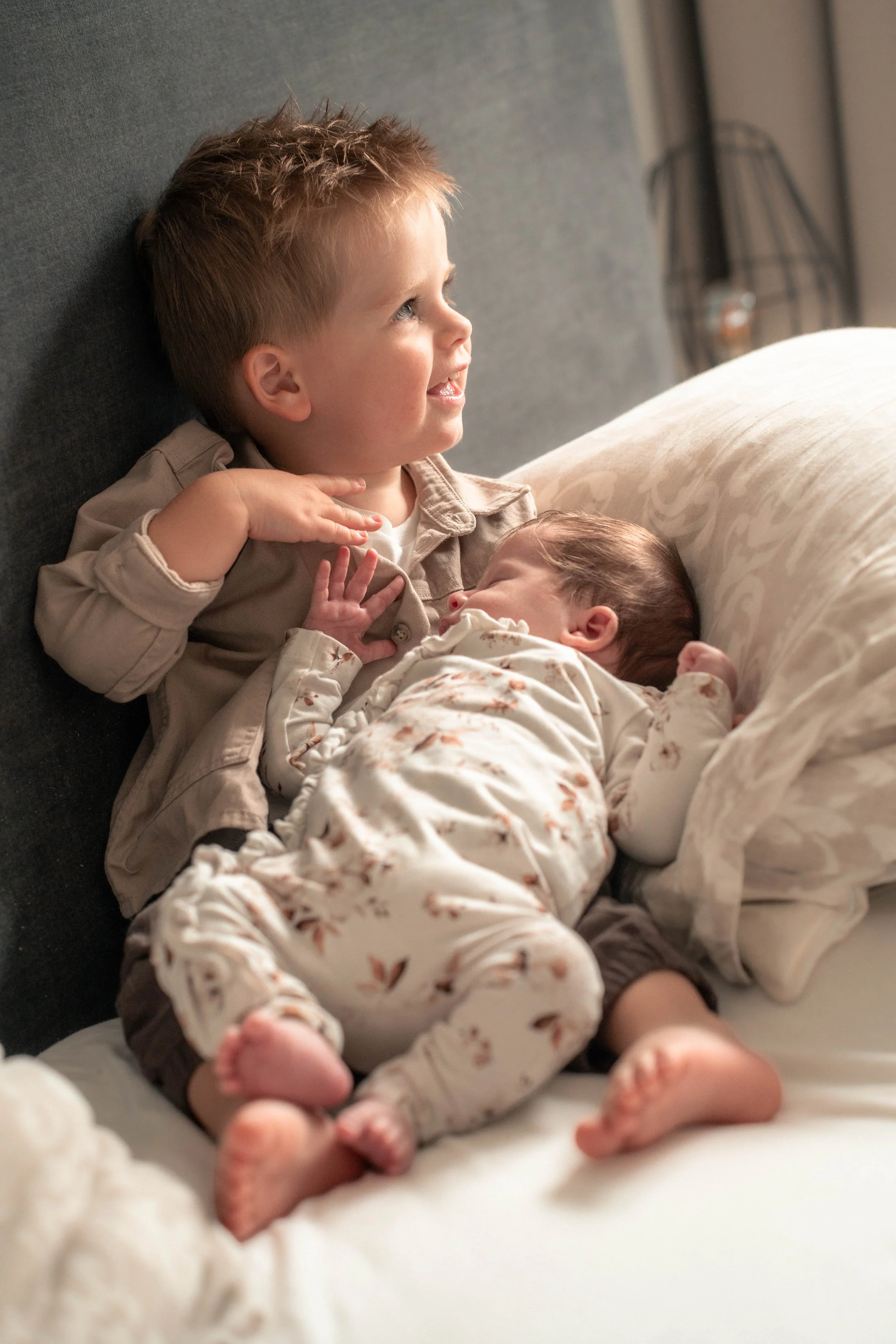 Een jonge jongen met blond haar ligt op zijn zij op een bankje, glimlachend, terwijl hij een pasgeboren baby vasthoudt.