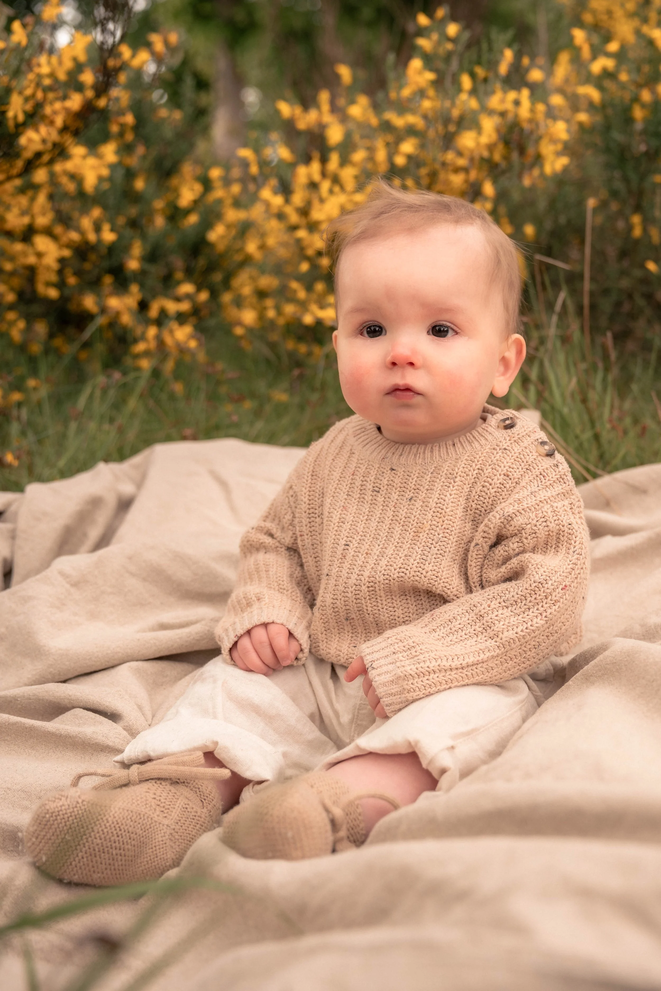 Een baby zit buiten op een beige deken, omgeven door groene en gele planten en struiken met gele bloemen, op een herfstachtige dag.