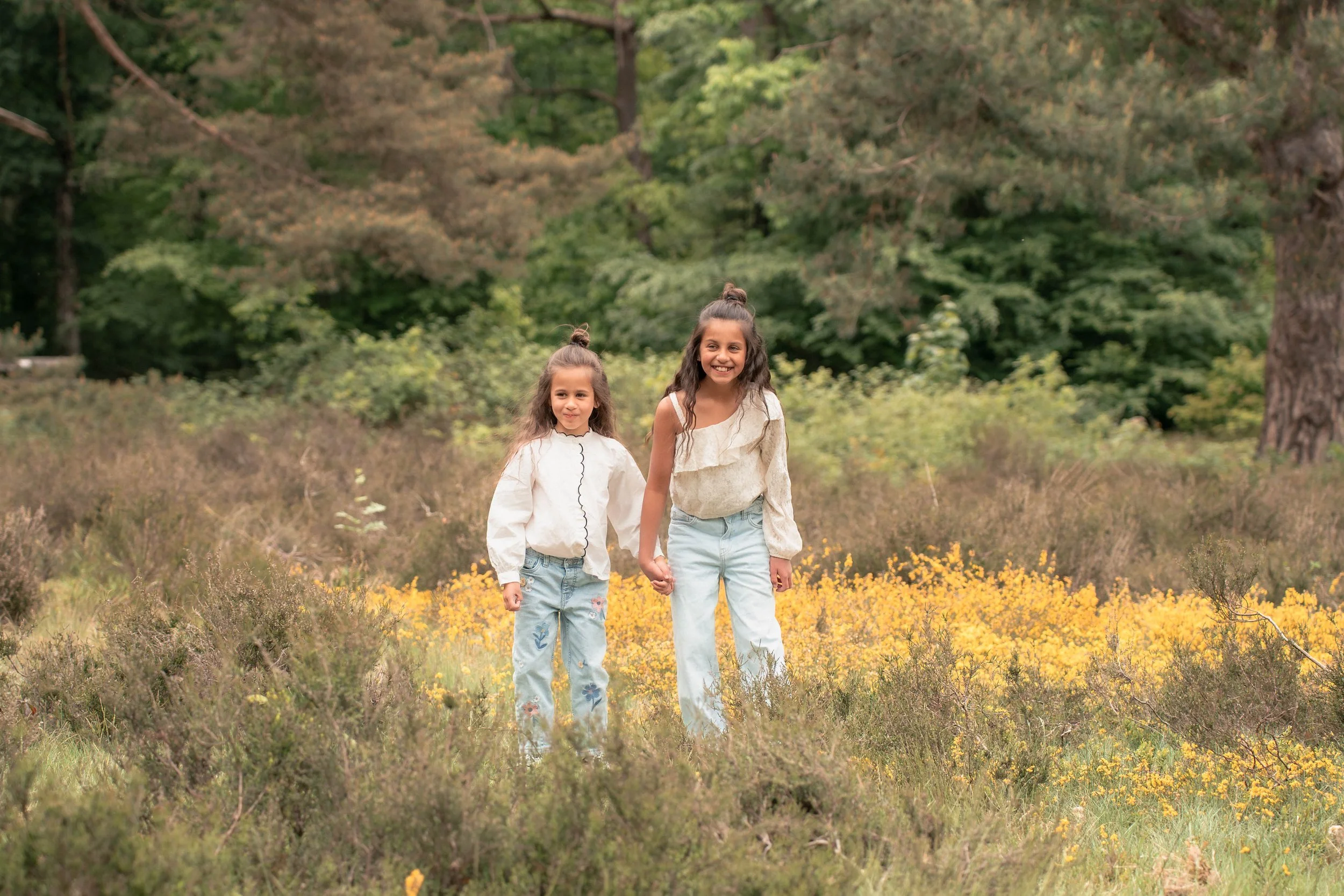 Twee meisjes wandelen hand in hand door een veld met gele bloemen, omgeven door groene bomen.