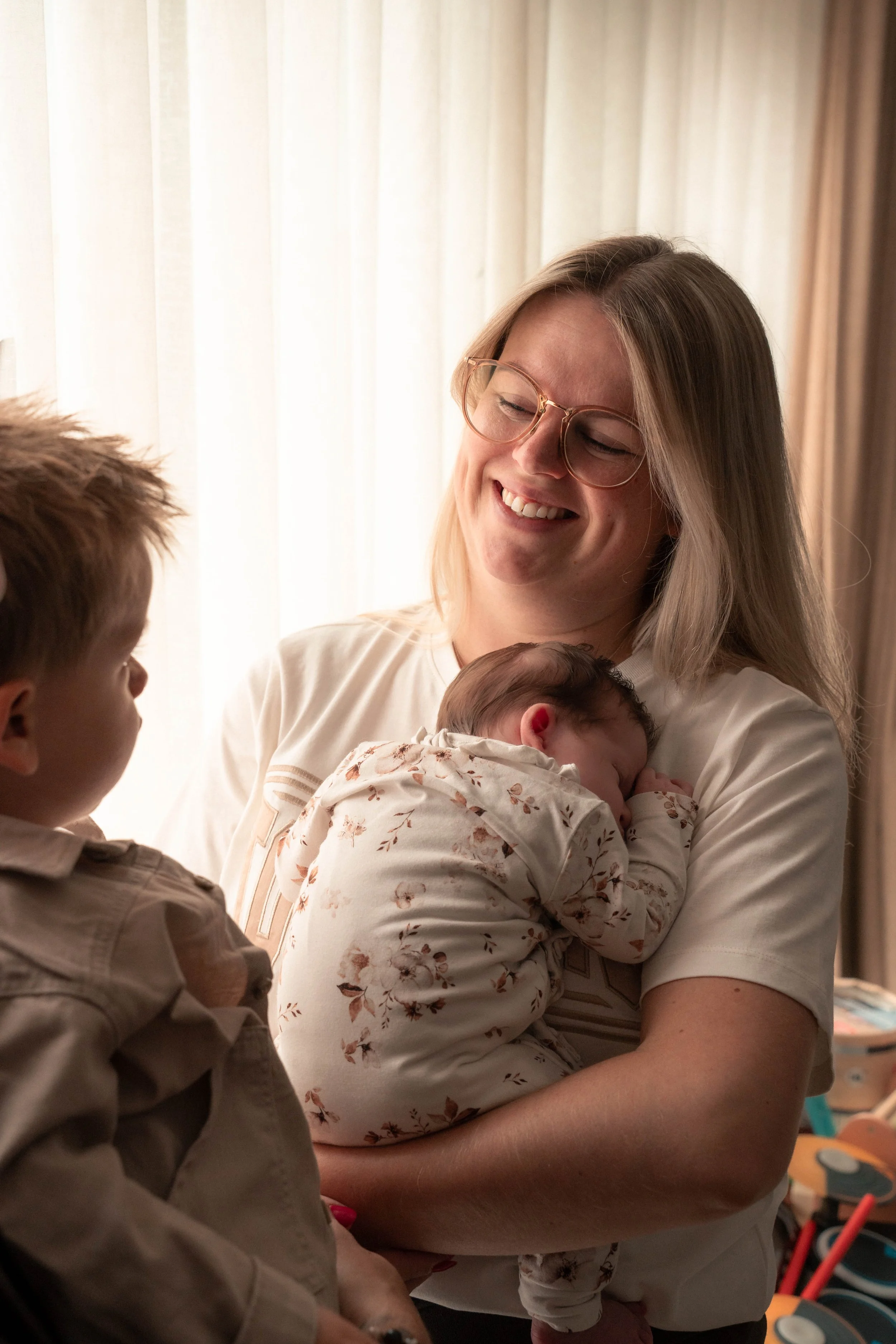 Een vrouw die een pasgeboren baby vasthoudt en glimlacht, terwijl er twee jonge kinderen in de buurt zijn.