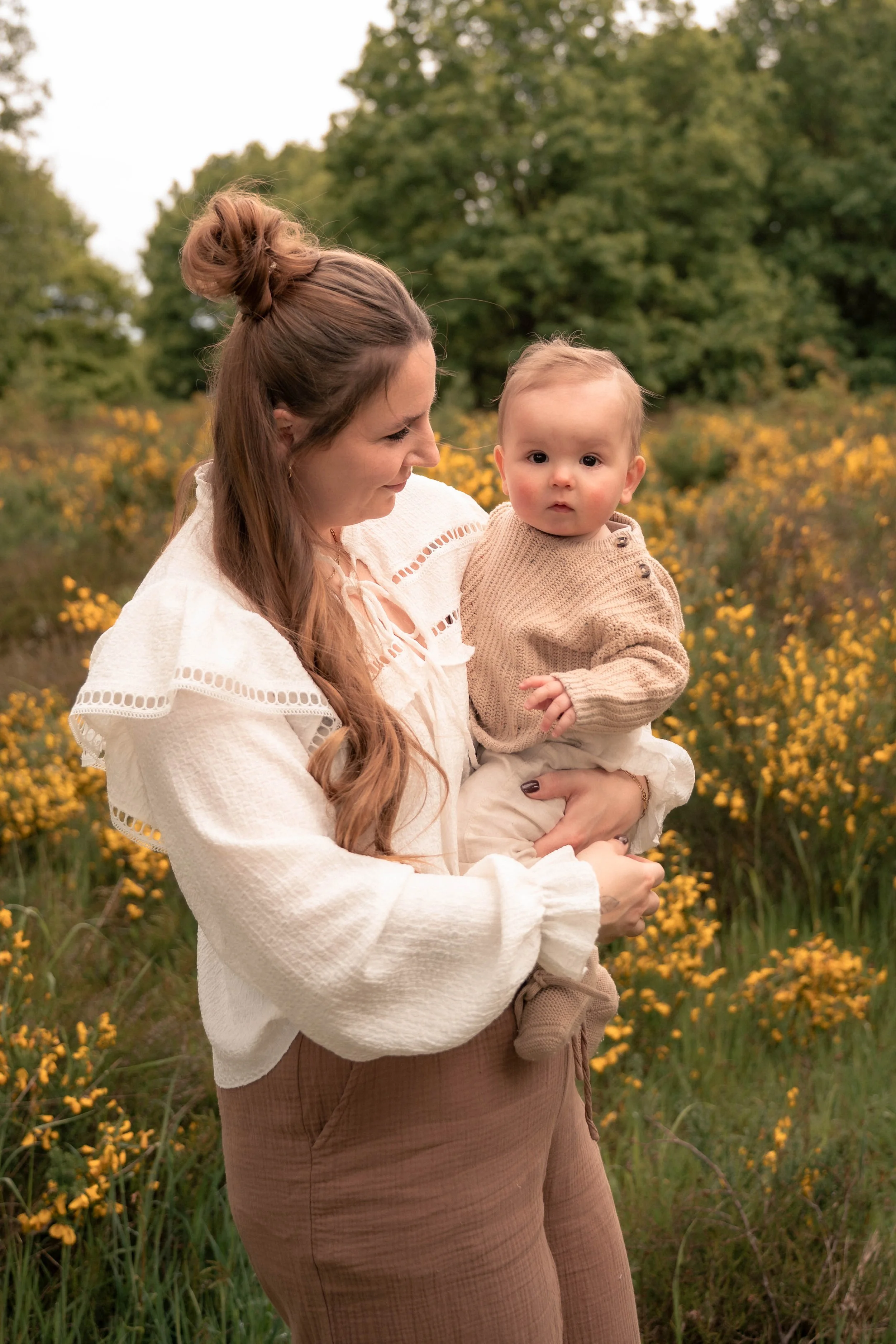 Vrouw houdt een baby in een oranje-bruine trui in de natuur met gele bloemen en groene bomen op de achtergrond.
