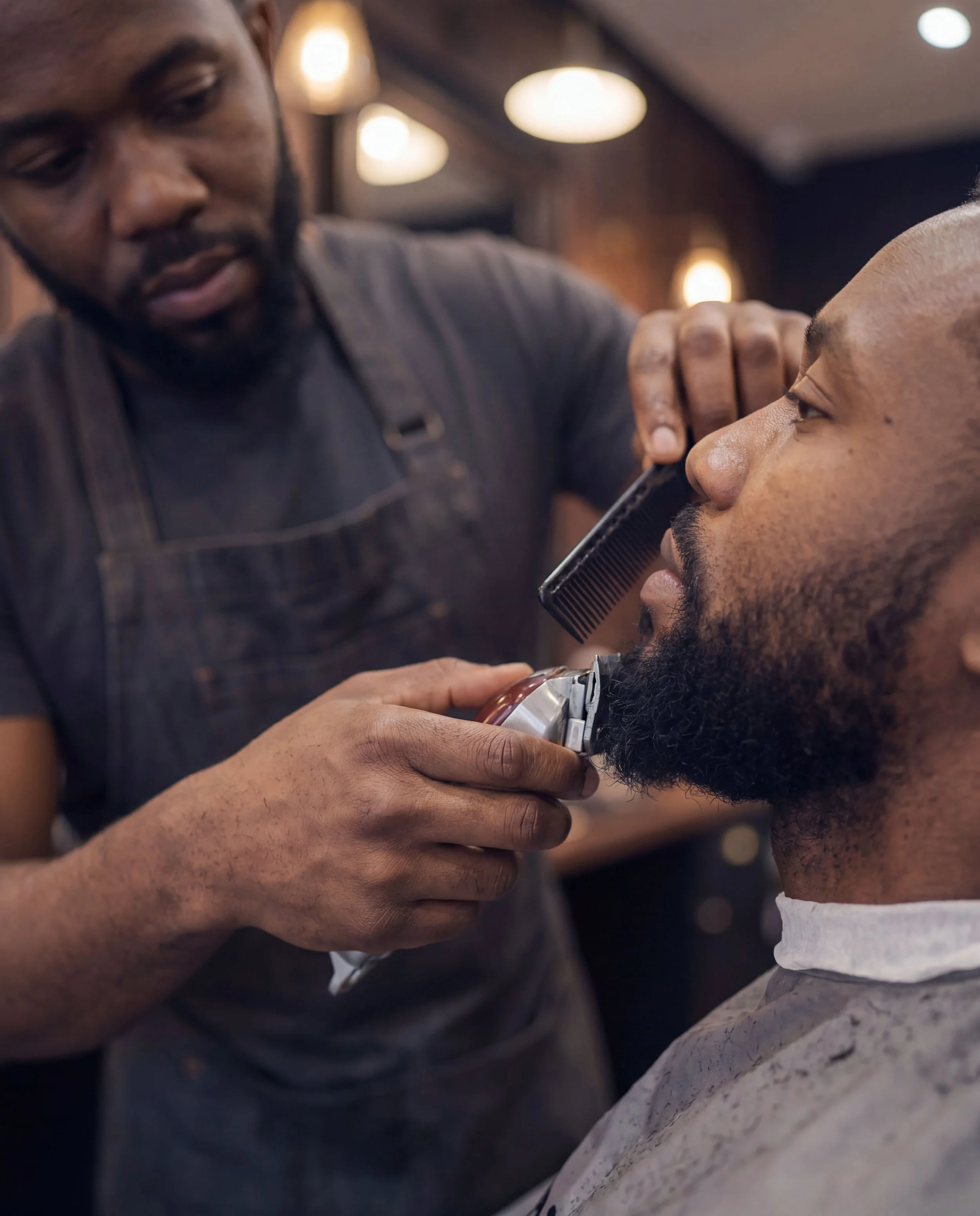 A barber trims a seated client's beard with clippers while holding a comb steady against his jaw, warm pendant lighting overhead.