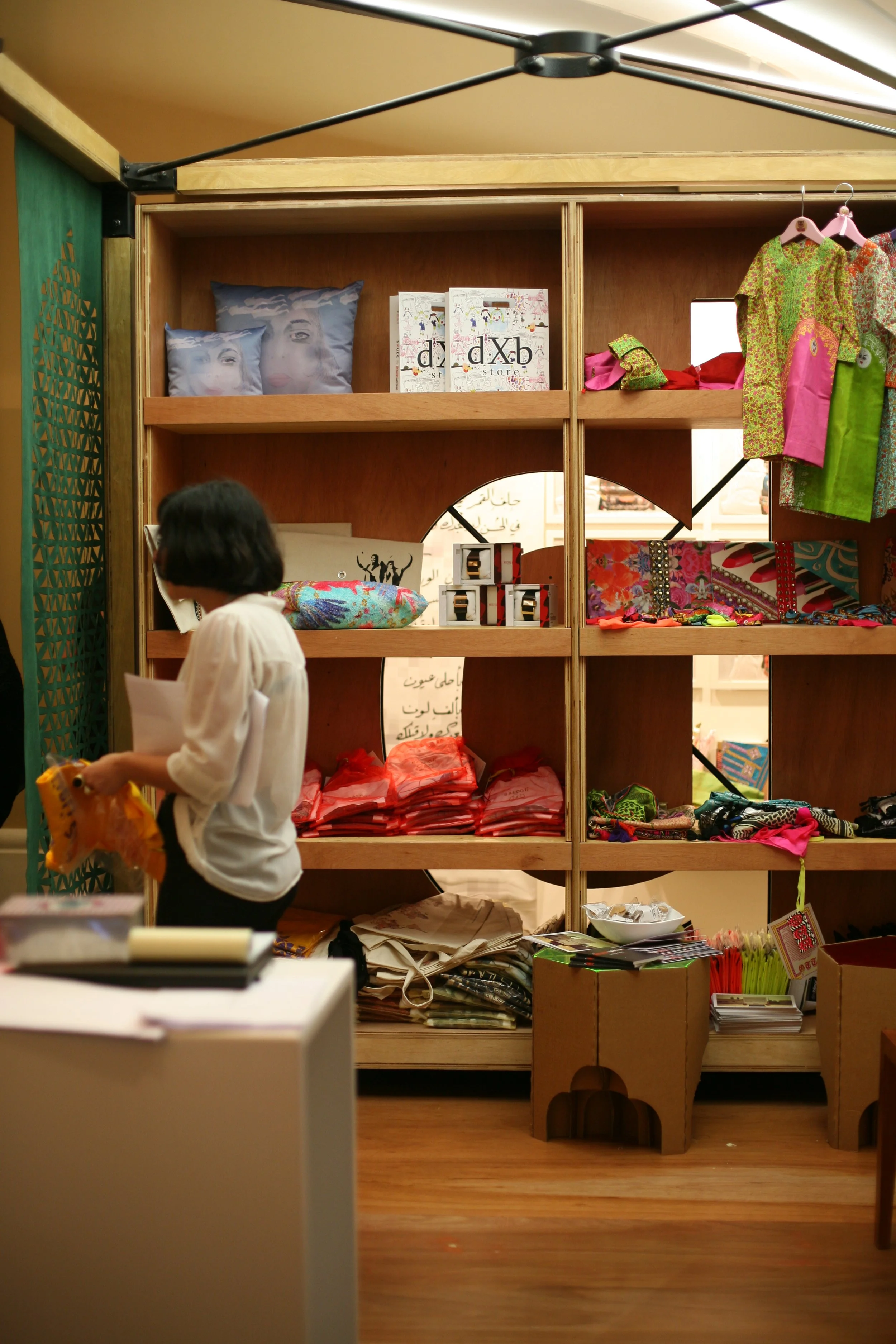 A woman looking at items on a table inside a store or market, with shelves containing colorful pillows, bags, clothing, and accessories behind her.