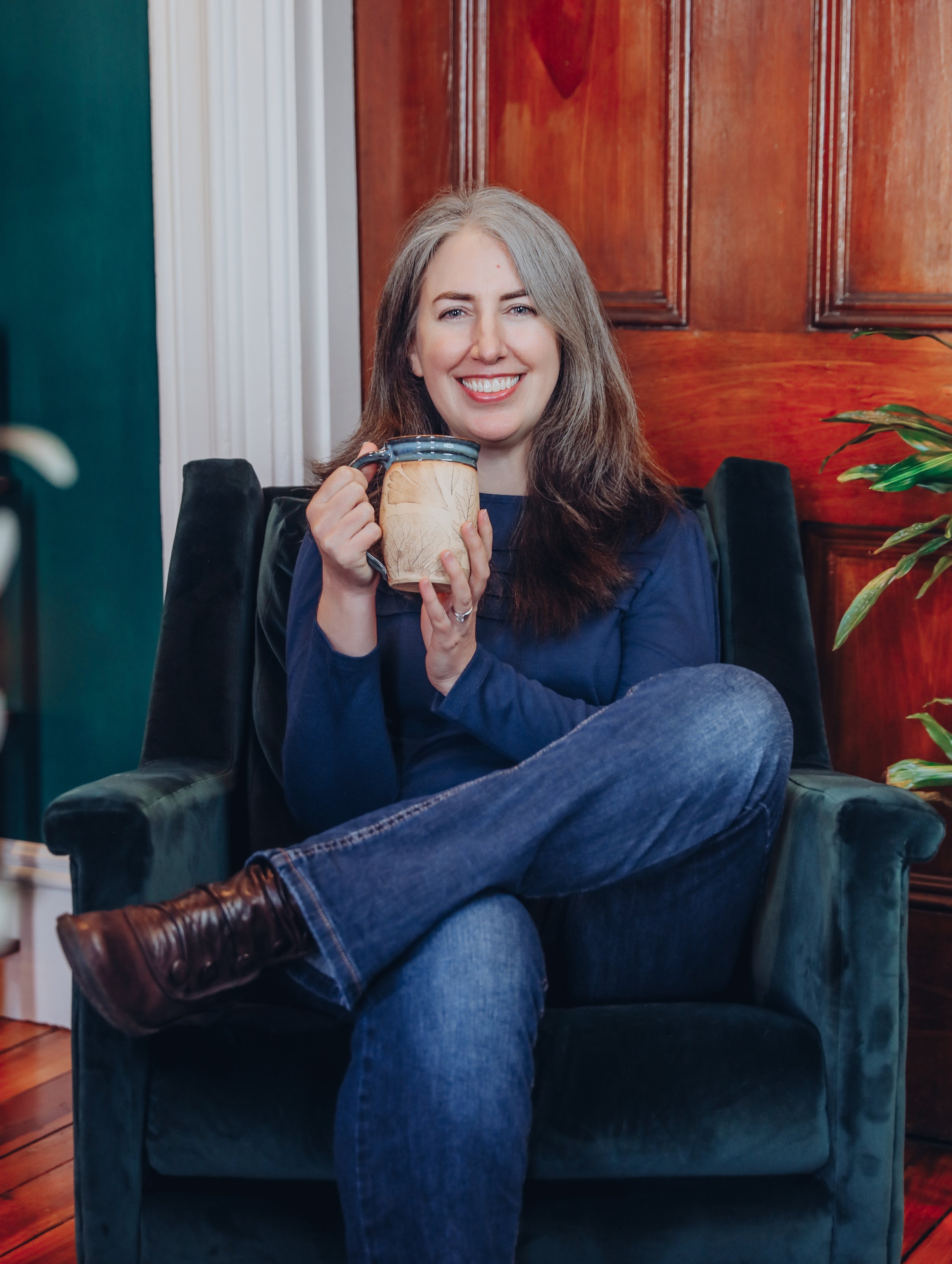St. Louis doula Kathleen Robbins sits in a chair holding a mug. Kathleen offers postpartum care to new moms.