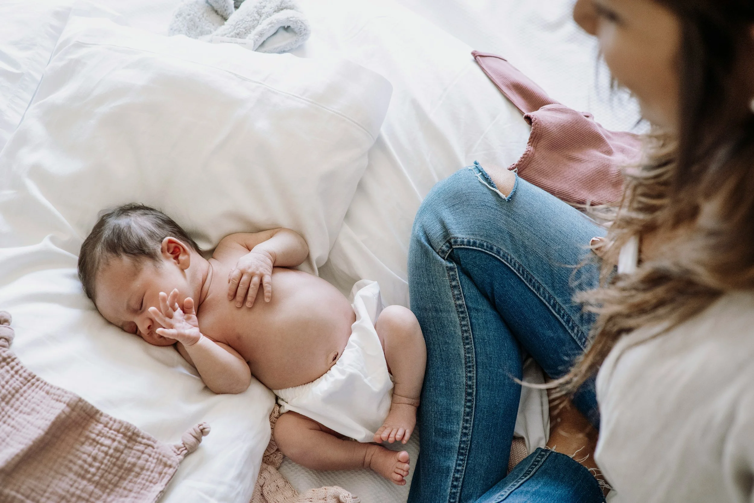 A baby lying on a bed with a blanket, next to a woman sitting on the bed, with curly hair and wearing ripped jeans and a pink jacket.