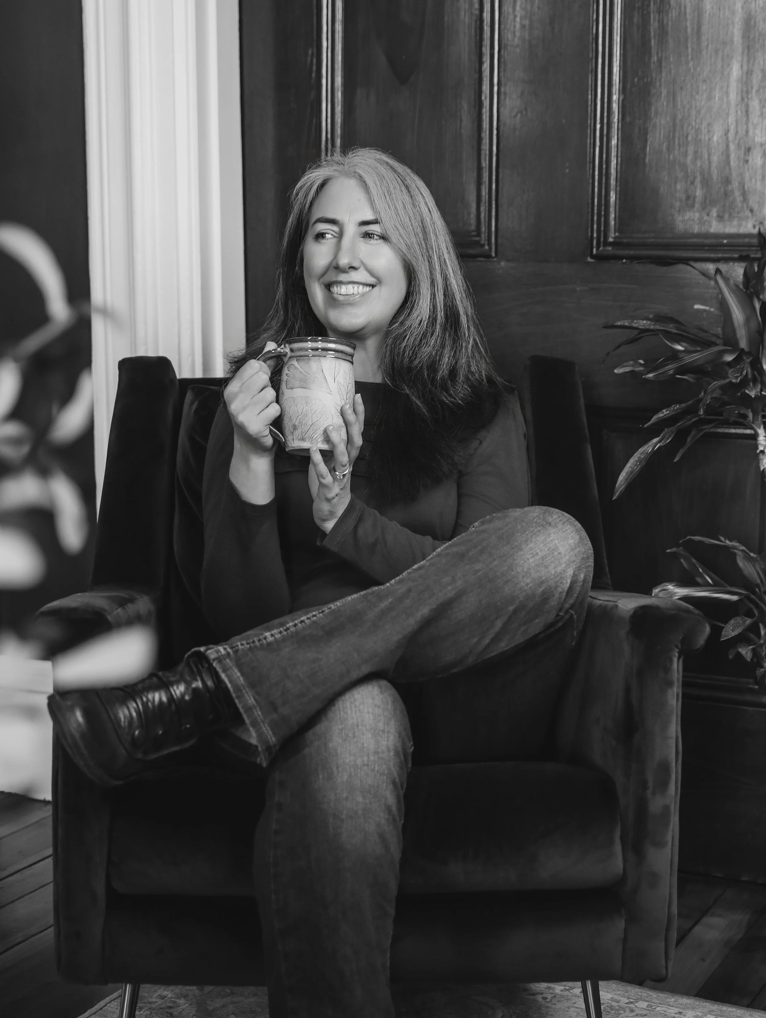 A woman with long hair smiling, holding a ceramic mug, sitting on a chair in a room with wooden walls and a plant nearby, in black and white.