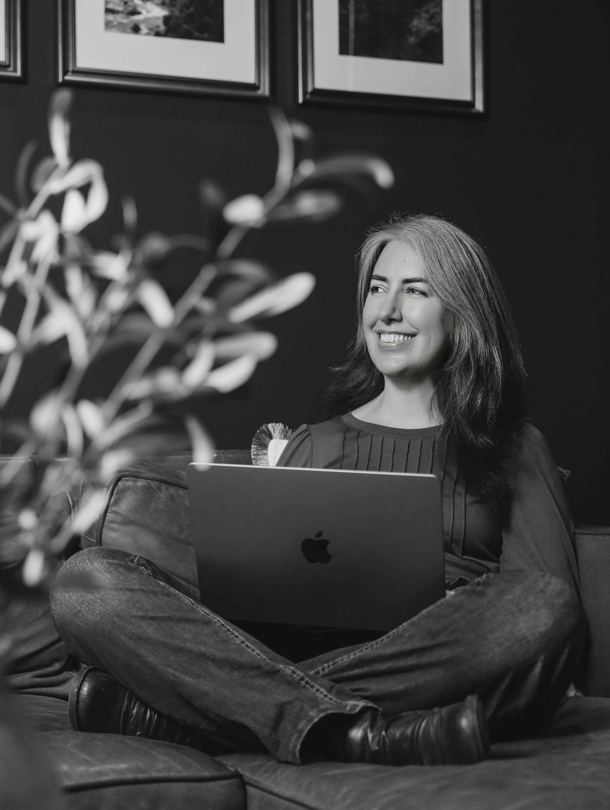 A woman with long hair sitting on a couch with a laptop in her lap, smiling and looking to her right. There are framed pictures on the wall behind her and a plant partially visible in the foreground.