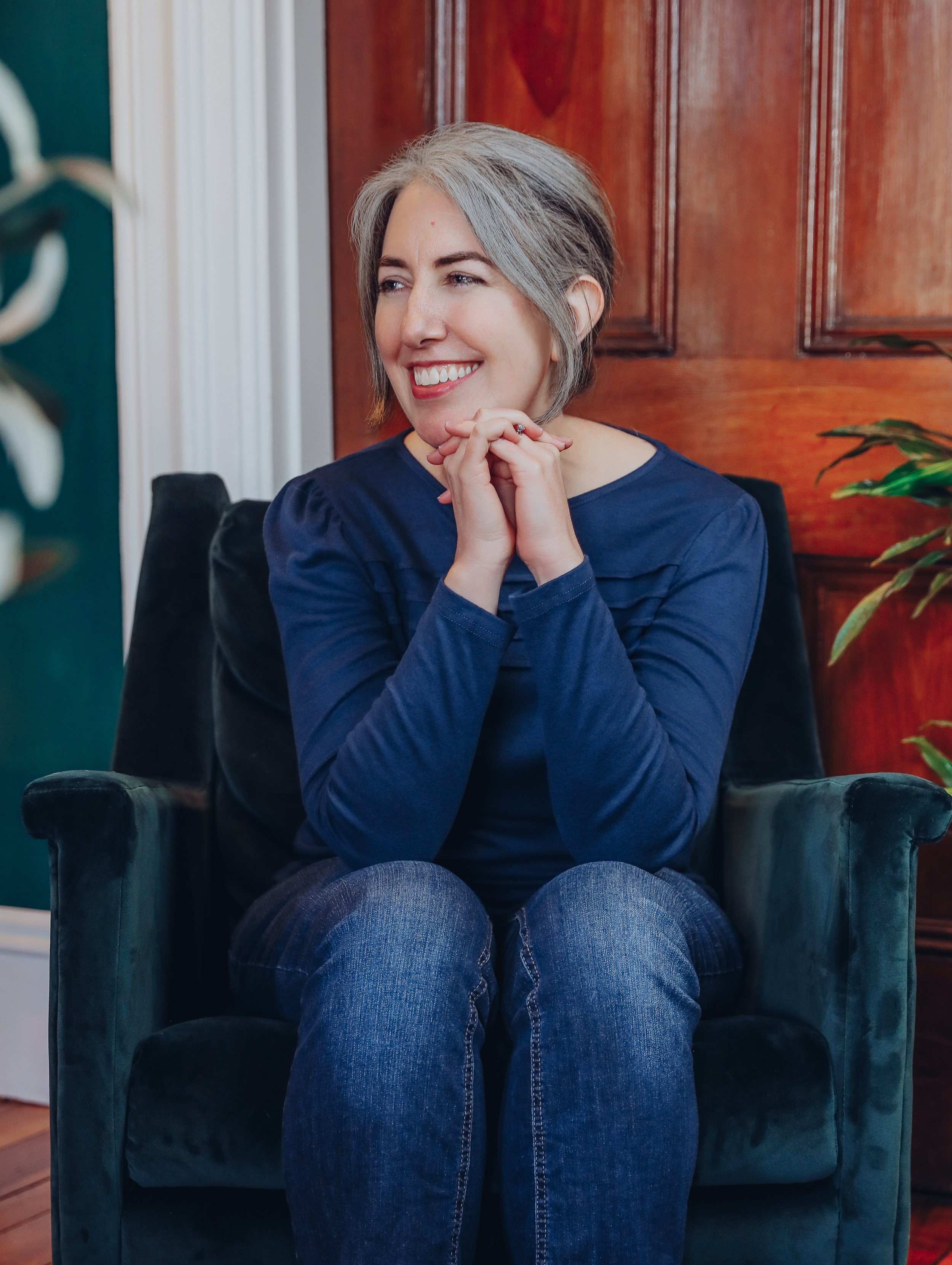 Woman with short gray hair smiling, seated on a dark green armchair, with a wooden wall behind her and a plant visible on the right.