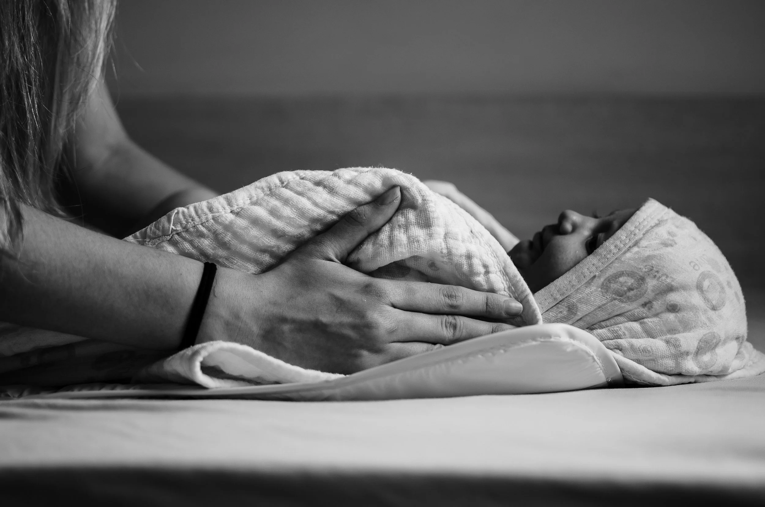 Black and white photo of a person’s hands holding a blanket or cloth, with part of their arm and hair visible on the left side. The setting appears to be indoors with a flat surface underneath.