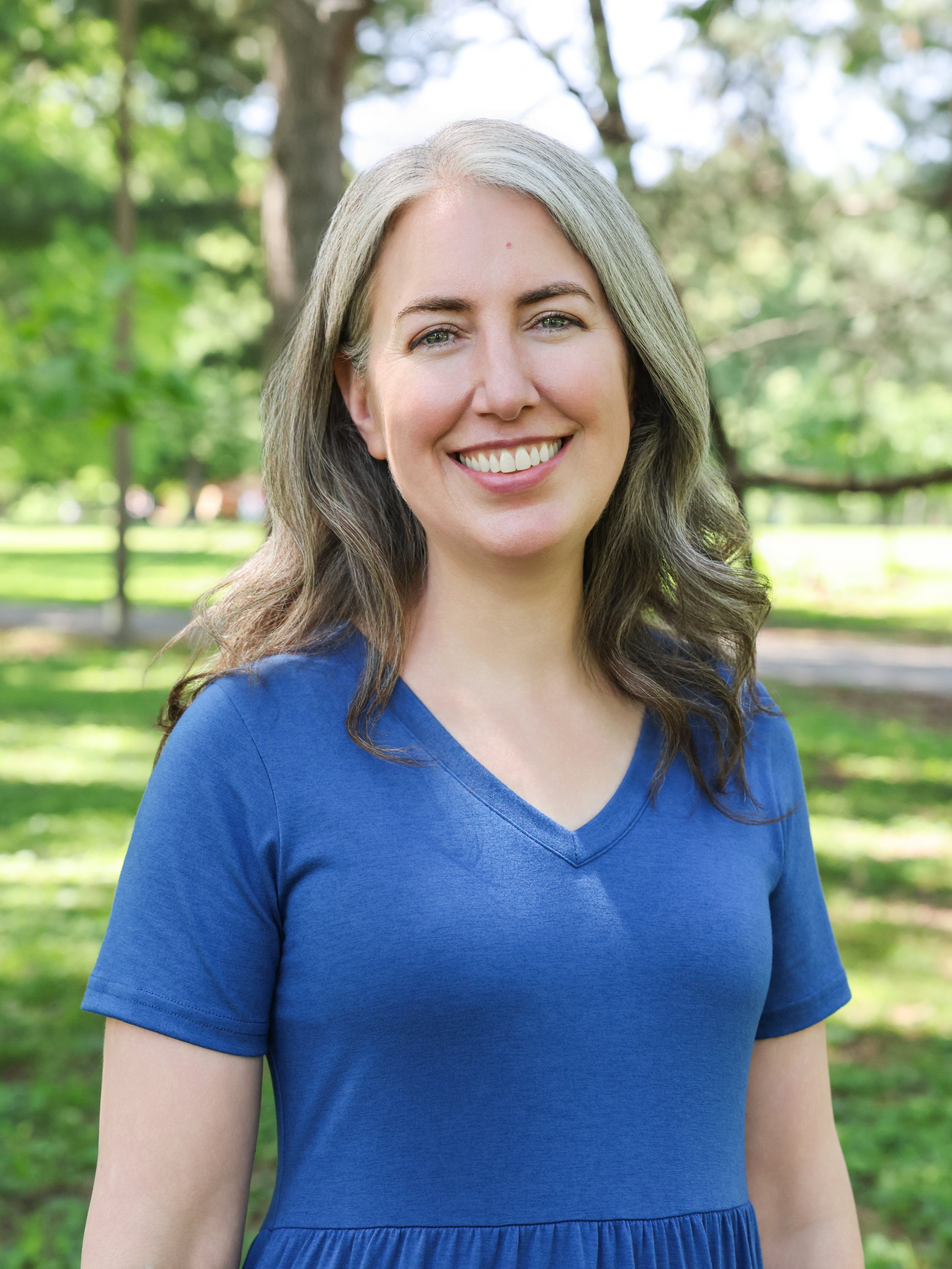 St. Louis postpartum doula Kathleen Robbins stands in a park wearing a blue dress.
