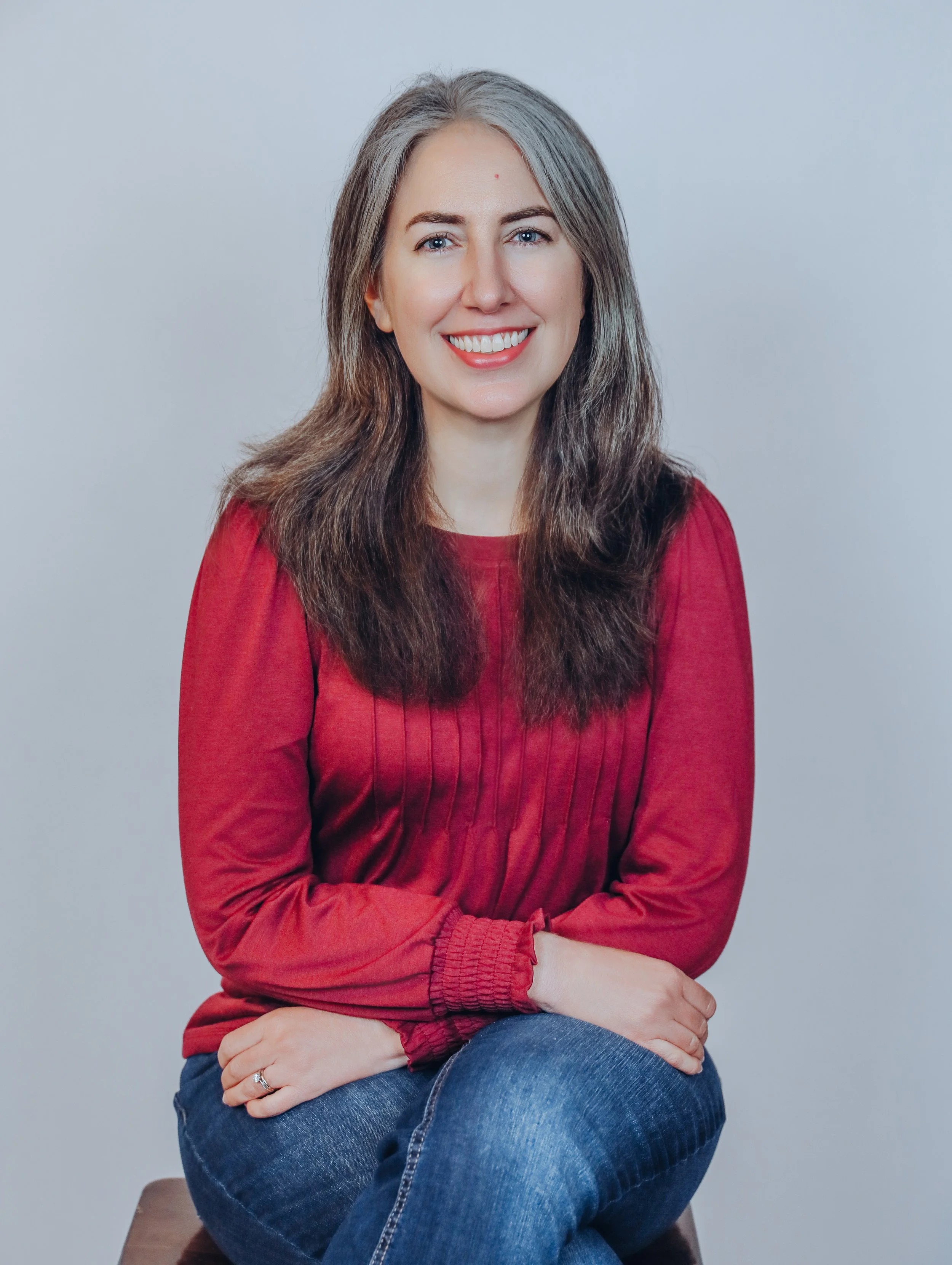 Portrait of a woman with gray hair wearing a red top and blue jeans, sitting with crossed arms against a plain light gray background.