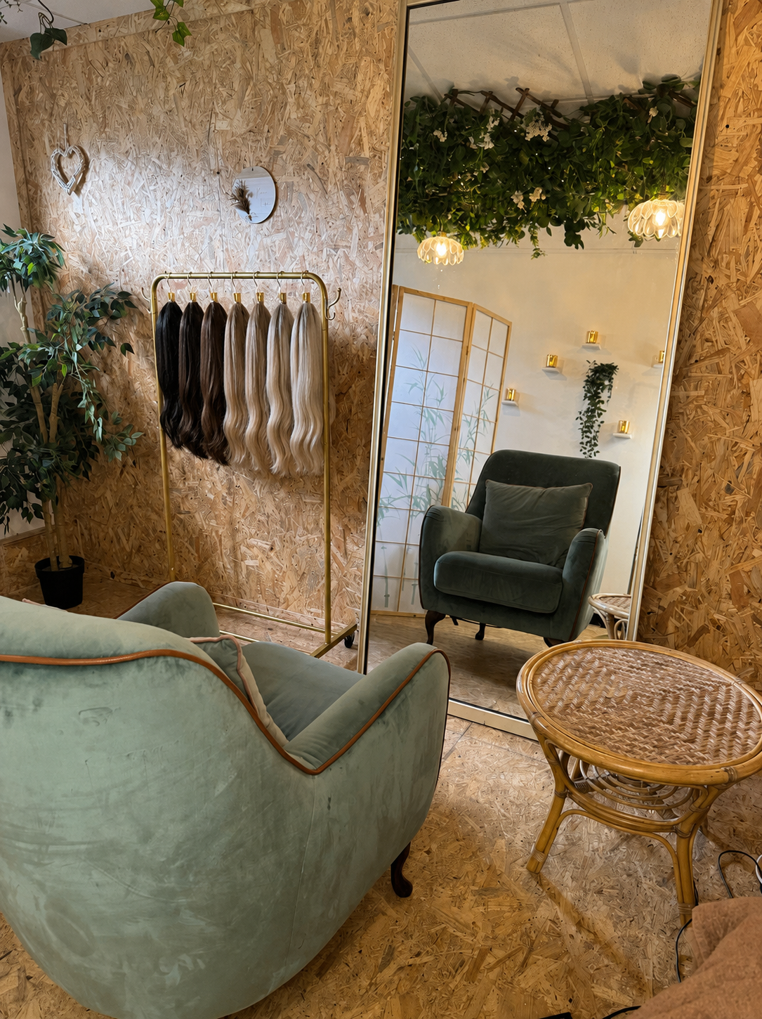 A cozy room with a large mirror reflecting a green armchair, a woven rattan side table, and a decorative ceiling with greenery and hanging lights. On the wall, there are some small shelves holding candles and plants, and a wooden wall with hearts and a mirror with hair extensions hanging on a golden clothing rack.