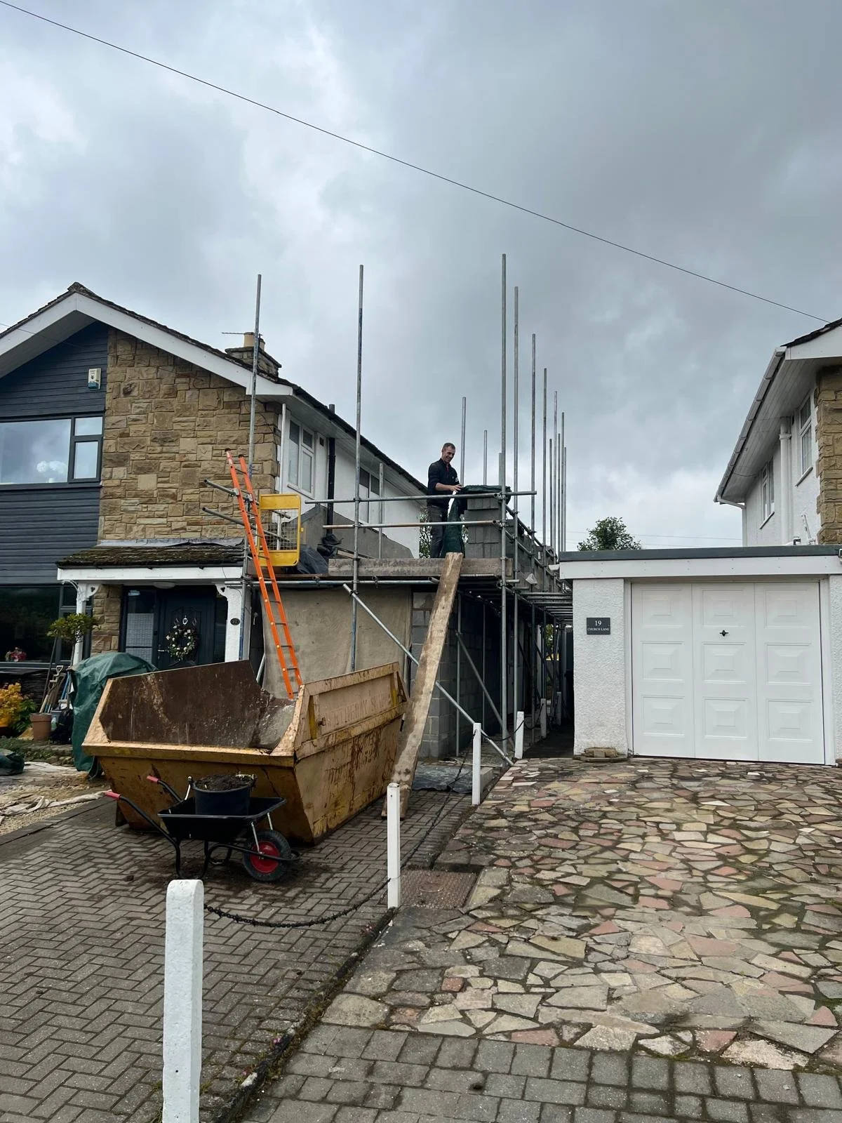A house under construction with scaffolding, a person working on the upper level, and construction materials including a wheelbarrow and a large rusty skip in the driveway. The scene is outdoors on a cloudy day.