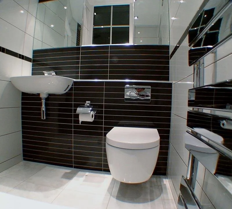 Modern bathroom with a white toilet, a small sink, a toilet paper holder, and a mirrored wall in the background.