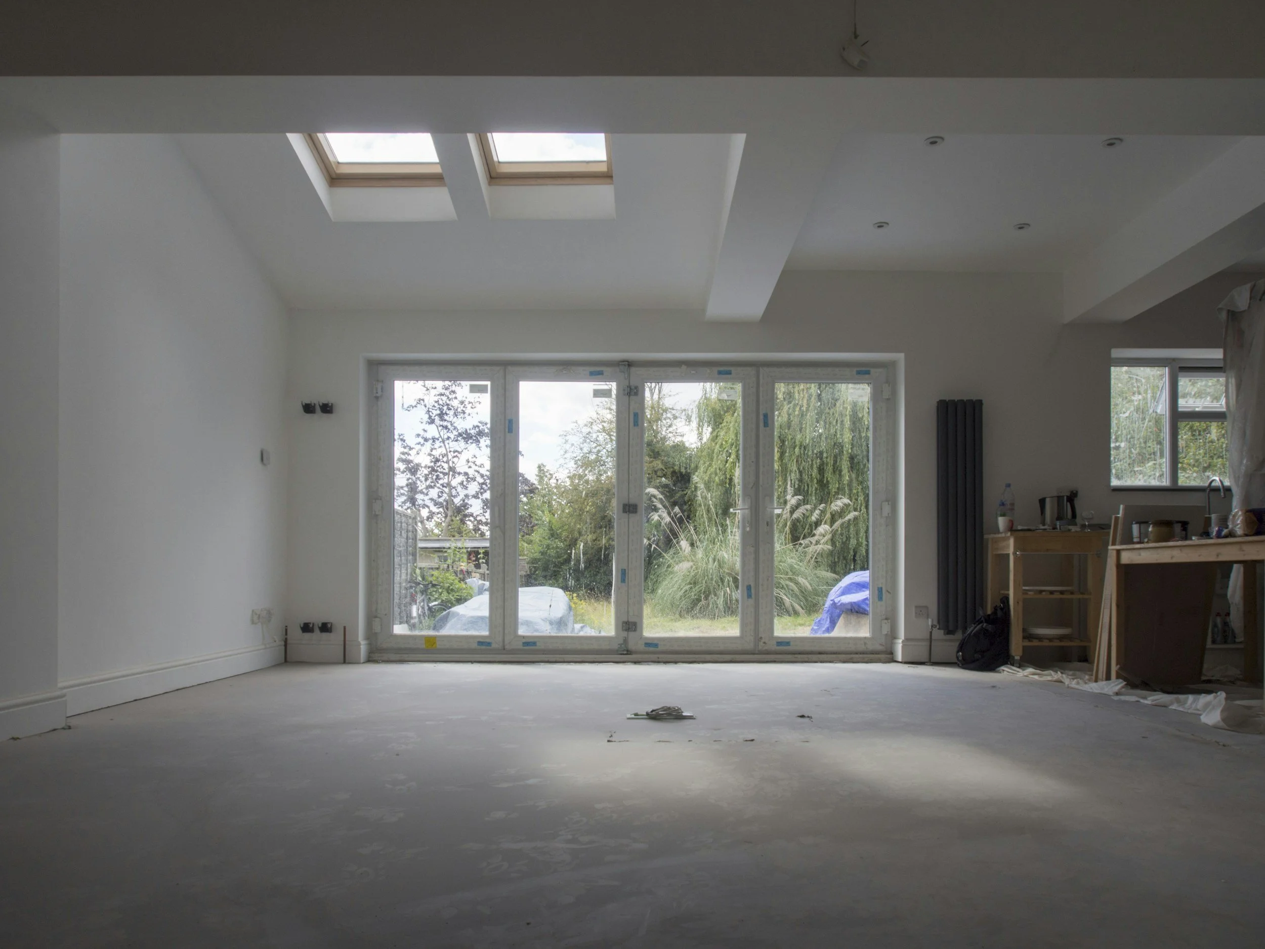 An unfinished room with large sliding glass doors overlooking a garden. Skylights in the ceiling allow natural light into the space, which is under construction or renovation.