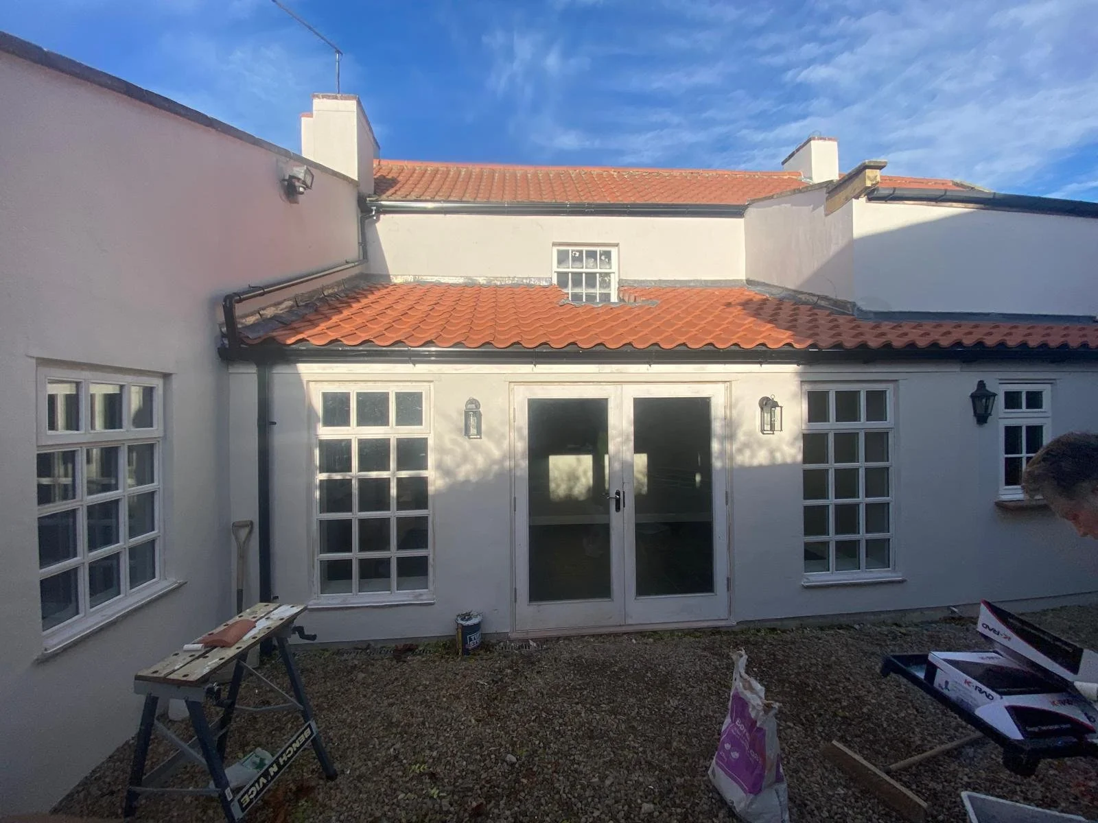Backyard patio area with white house walls, multiple windows, and a glass door, with a red tile roof and a blue sky overhead. There are tools and a bag of materials on the ground and work tables.