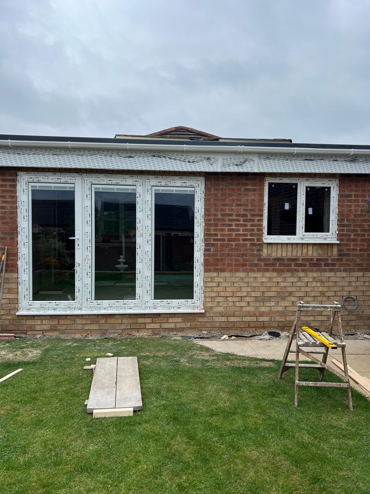 The back exterior of a brick house under renovation with newly installed white-framed windows and a sliding glass door. There are construction tools and materials on the lawn and a patch of grass in front.
