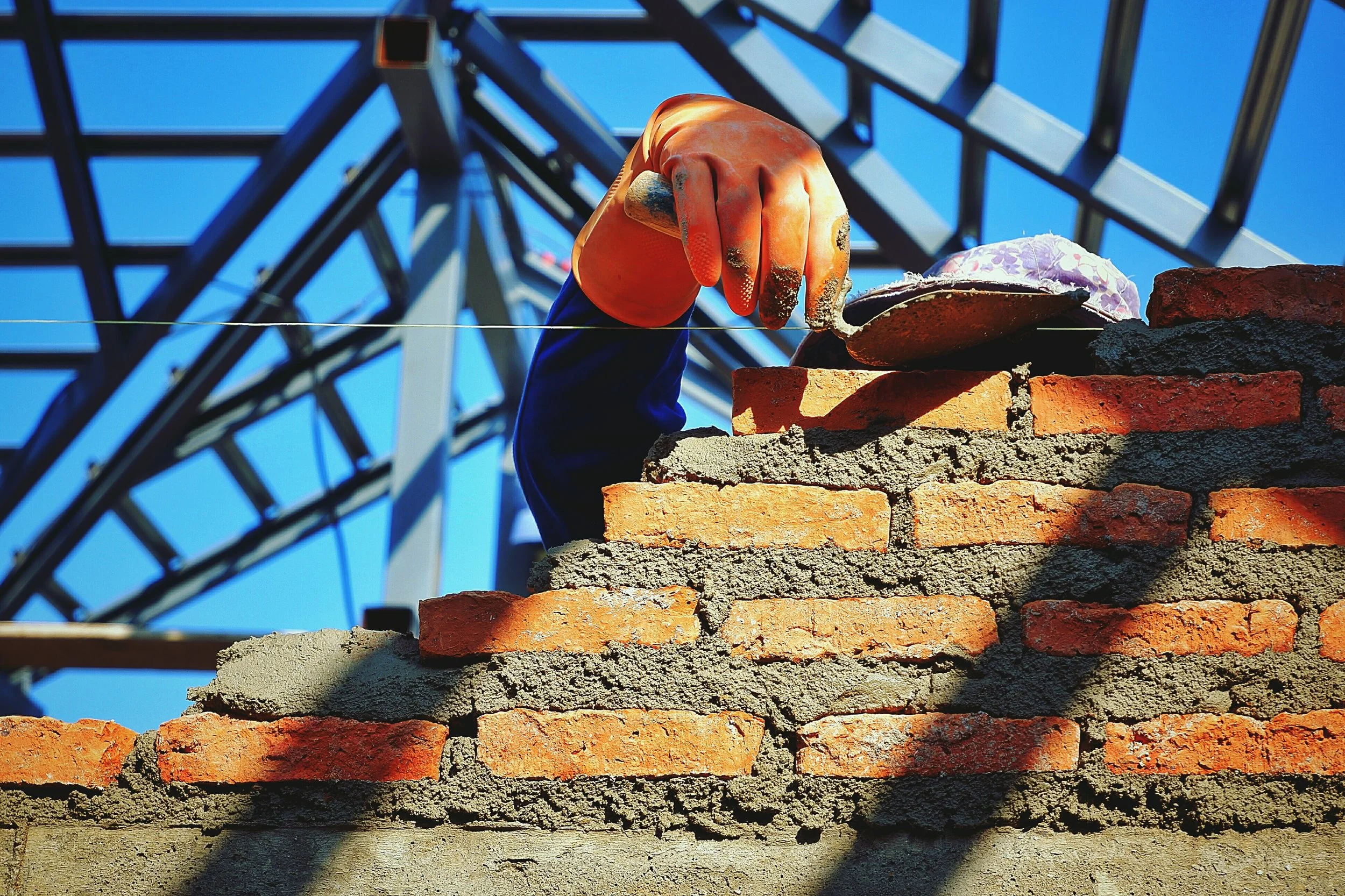 Close-up of a worker's hand wearing an orange glove, laying bricks on a wall under construction, with a metal structure and blue sky in the background.