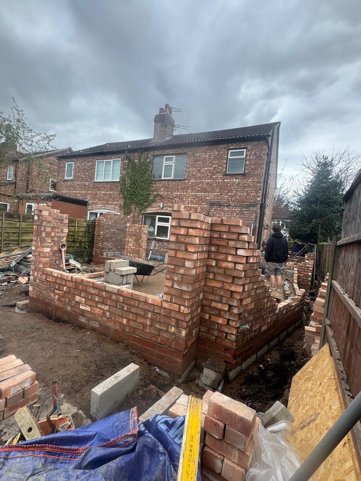 Backyard construction site with partially built brick wall, construction tools, and a man working, next to a brick house under cloudy sky.