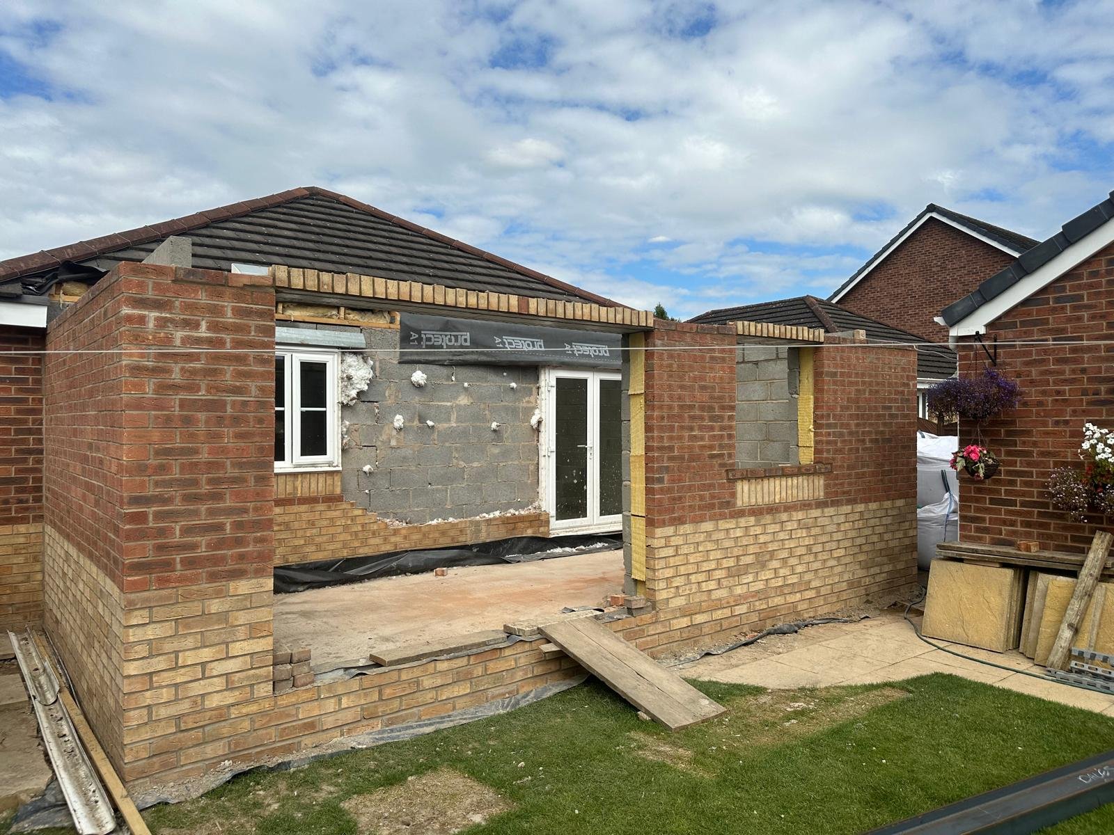 A house under construction with partially built brick exterior walls, a small window, and construction materials and tools on the ground.